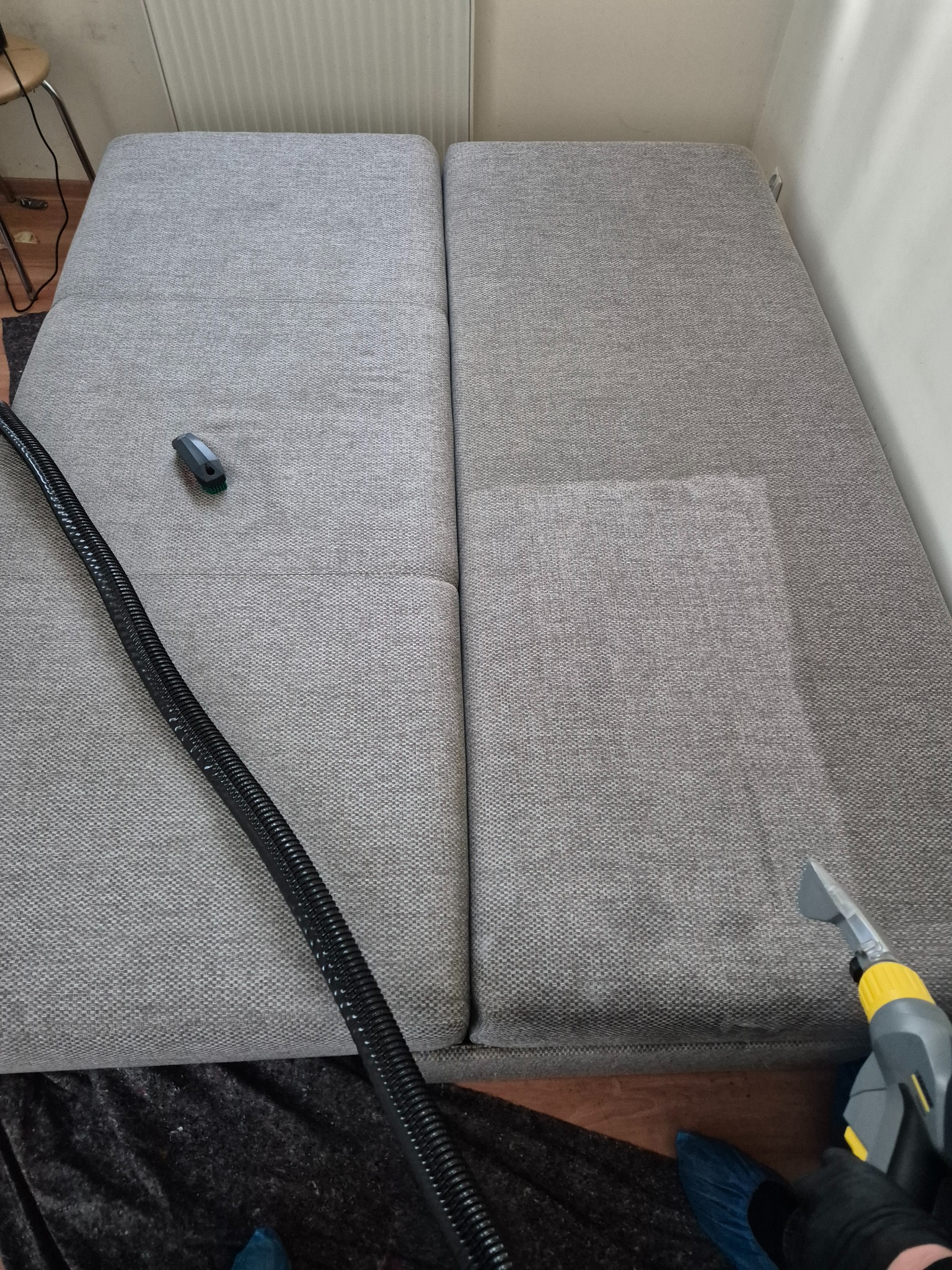 Szara kanapa modułowa, częściowo wyczyszczona, z widoczną różnicą w odcieniu, dysza odkurzacza piorącego z żółtymi elementami w prawym dolnym rogu, obok czarny wąż i mała szczotka.