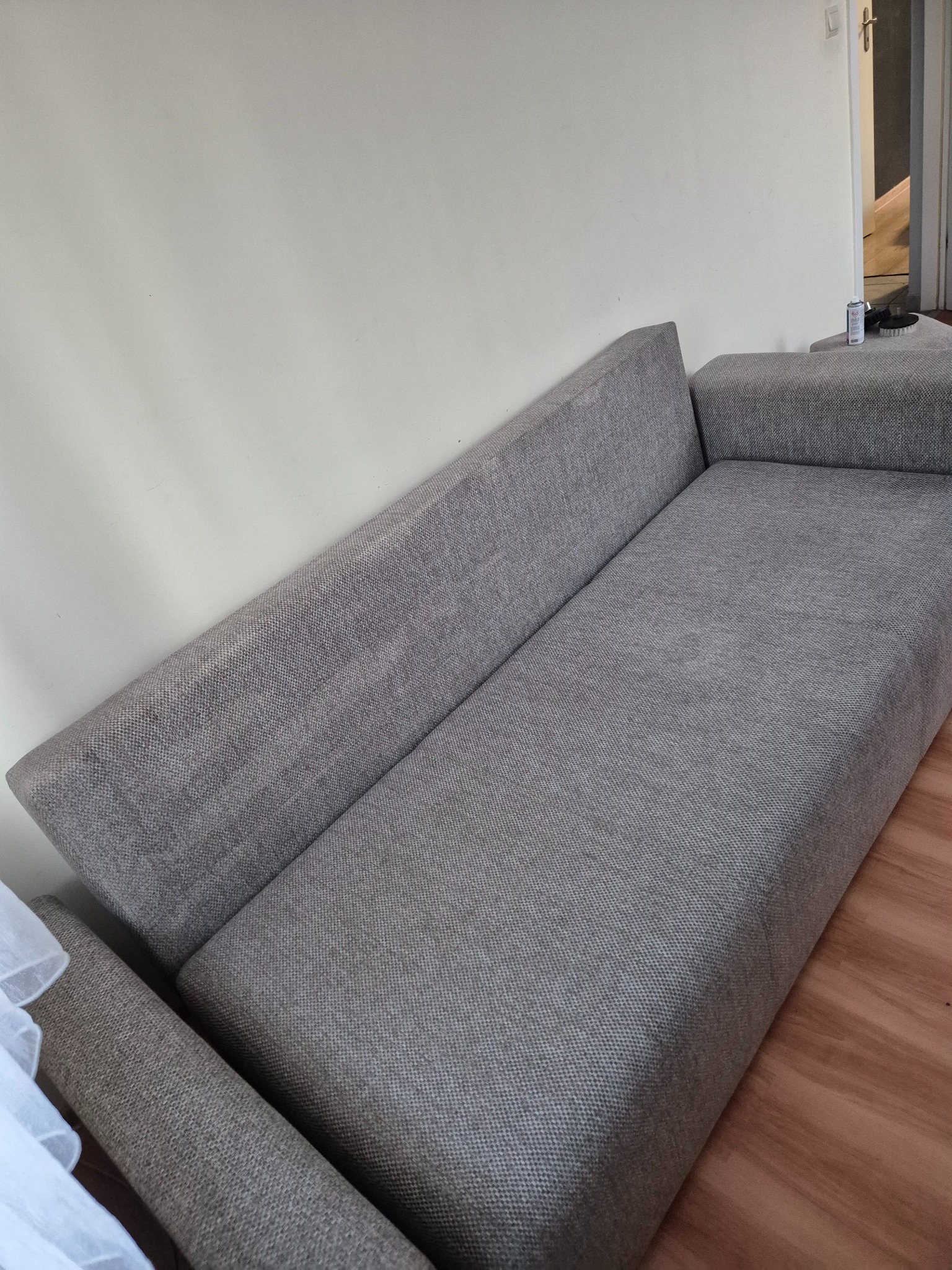 Szara kanapa z widocznym środkiem do czyszczenia tapicerki i szczotką na boku, ustawiona przy białej ścianie i drewnianej podłodze.