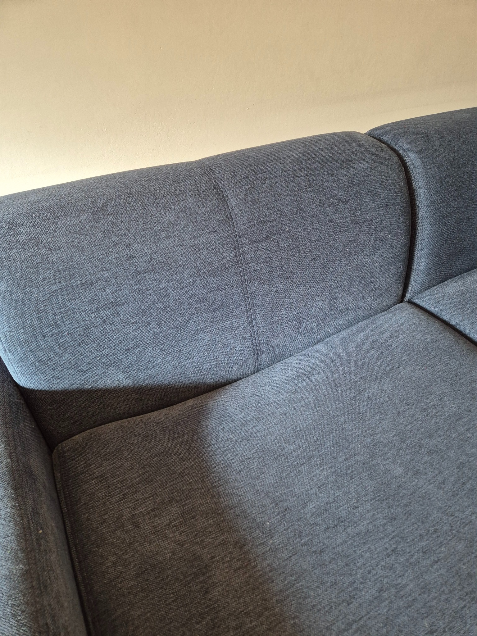 Fragment sofy z szarej tkaniny o wyraźnym splocie na tle beżowej ściany, widoczny szew łączący elementy oparcia.