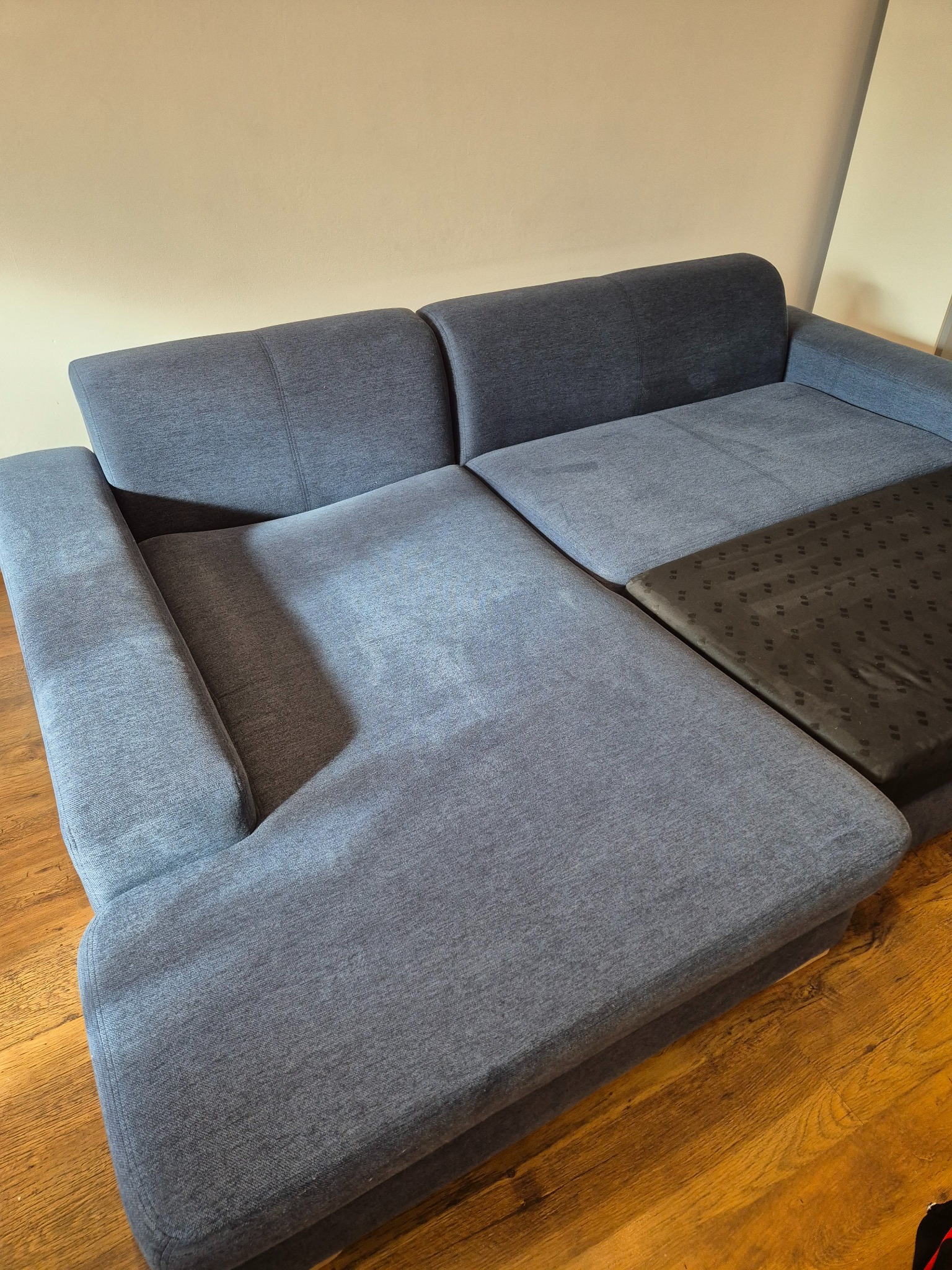 Szary narożnik z tkaniny o widocznej strukturze, częściowo rozłożony, z ciemniejszym, wzorzystym elementem, na drewnianej podłodze i tle beżowej ściany.