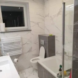 Nowoczesna łazienka z białymi płytkami imitującymi marmur, szarą obudową toalety i wanny, grzejnikiem drabinkowym oraz kabiną prysznicową ze szklanymi drzwiami.