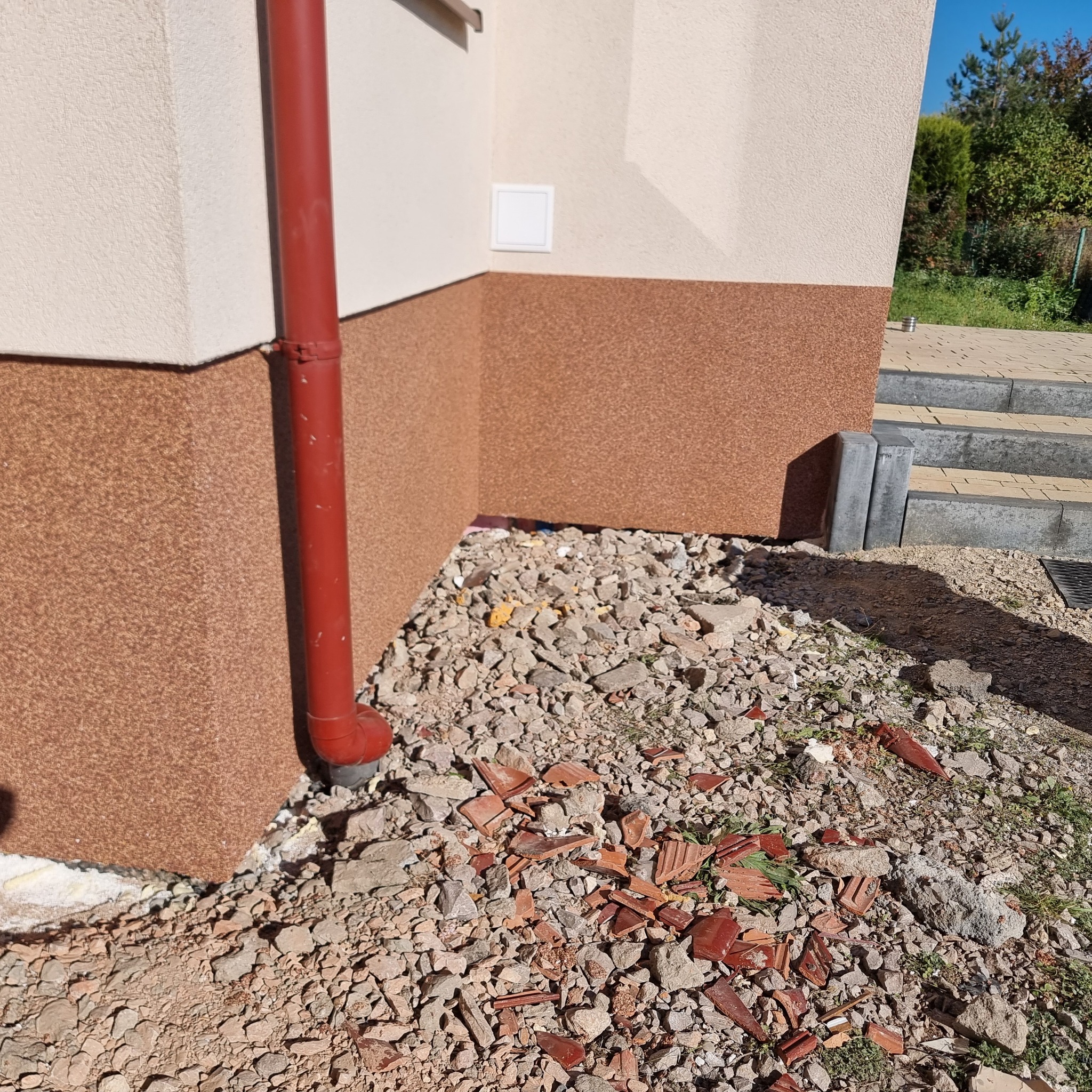 Fragment elewacji budynku z czerwoną rynną i fragmentami potłuczonych płytek ceramicznych u podstawy ściany.