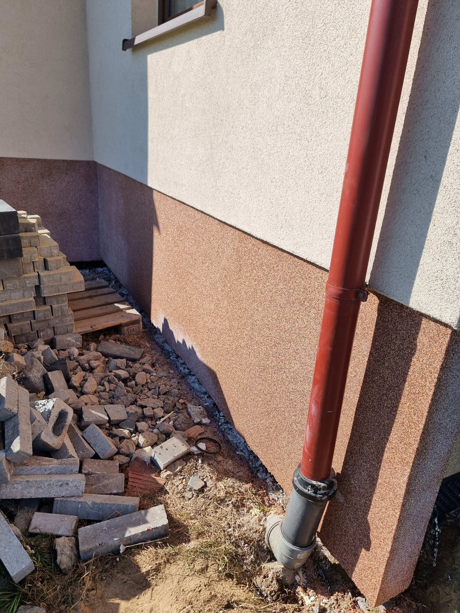 Przygotowania do ułożenia kostki brukowej przy ścianie budynku z zamontowaną rurą spustową, widoczny stos kostki na palecie i rozsypane elementy.