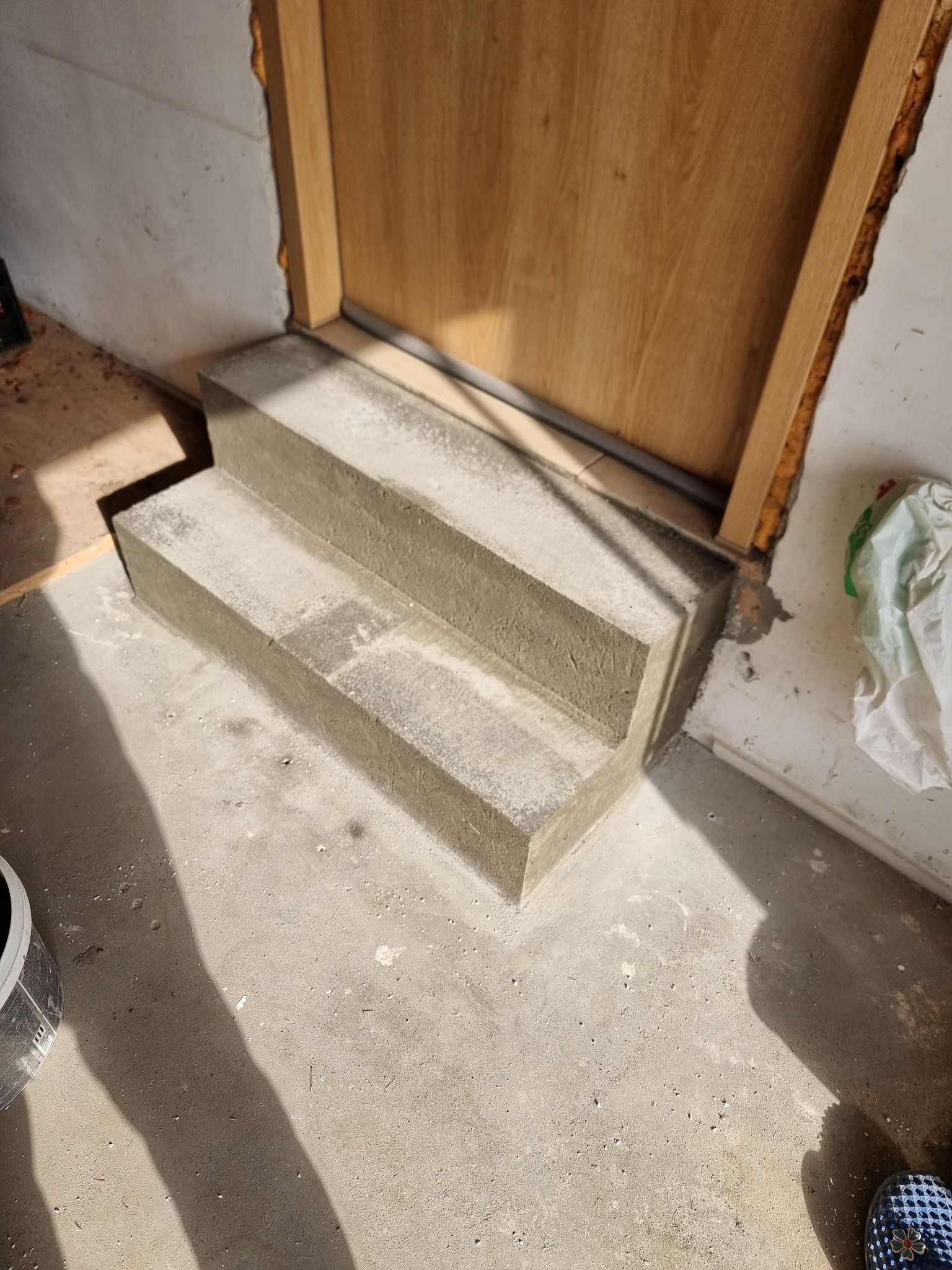 Betonowe schody wejściowe, świeżo wykonane, prowadzące do drewnianych drzwi, widoczne ślady zaprawy, surowe wykończenie wnętrza.