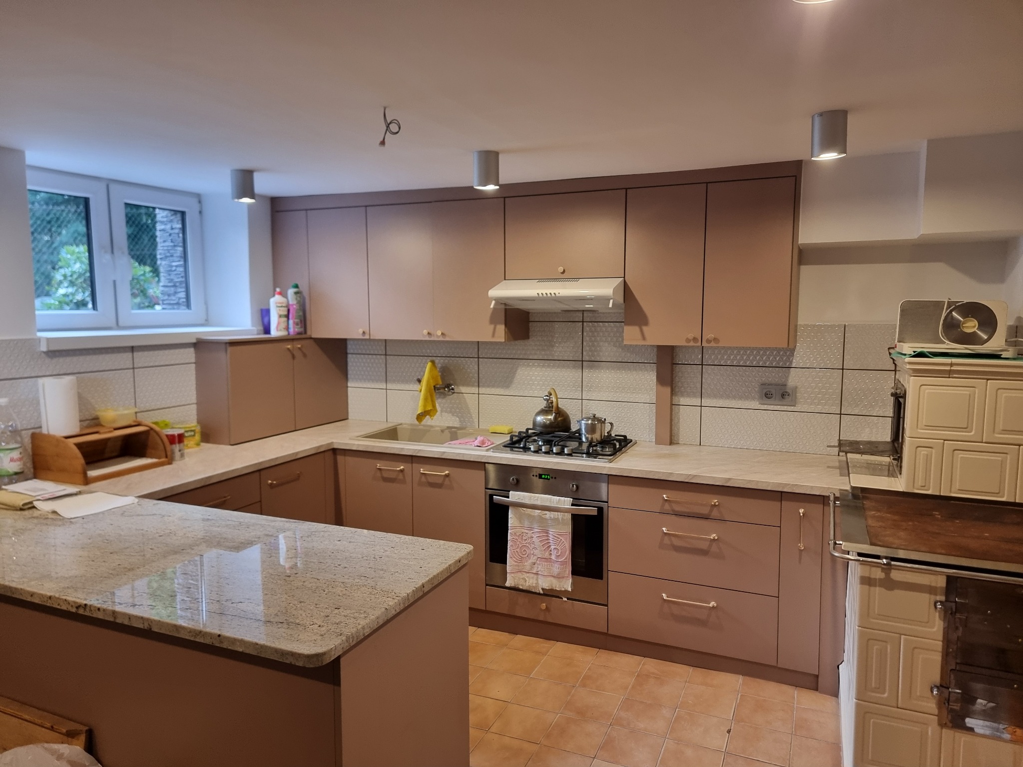 Jasna kuchnia z beżowymi szafkami, granitowym blatem i klasycznym piecem kaflowym, oświetlona punktowo, widok na okno z zielenią