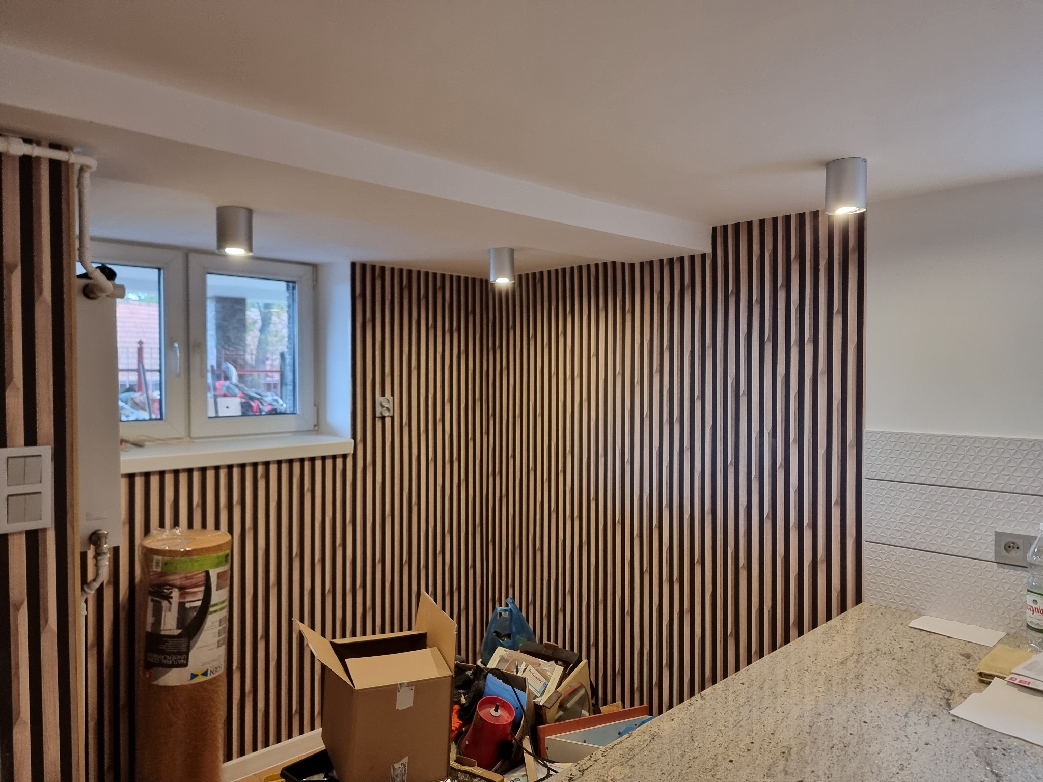 Montaż drewnianych paneli ściennych w pomieszczeniu z oknem i widocznymi puszkami instalacyjnymi, niedokończone prace, widoczny materiał izolacyjny w rolce i narzędzia w kartonie.