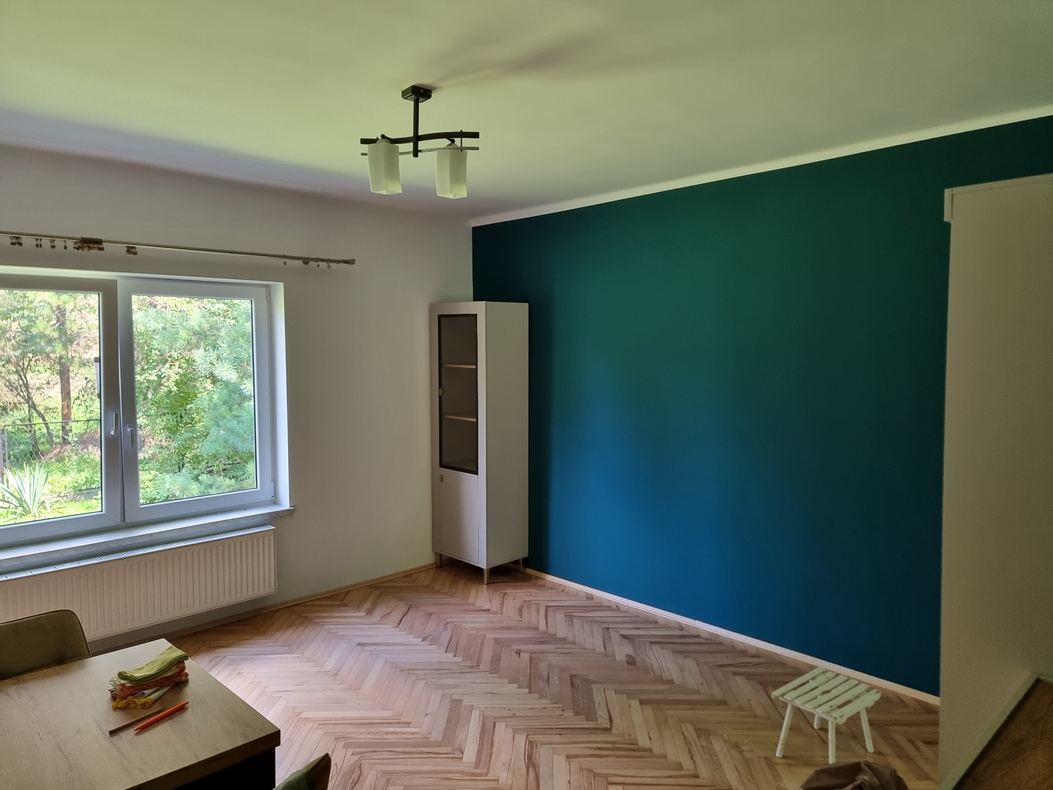 Pomalowane na ciemny turkus ściany w pokoju z parkietem w jodełkę, biała szafka narożna i okno z widokiem na zieleń, prosta lampa sufitowa, stół z materiałami i kredkami.