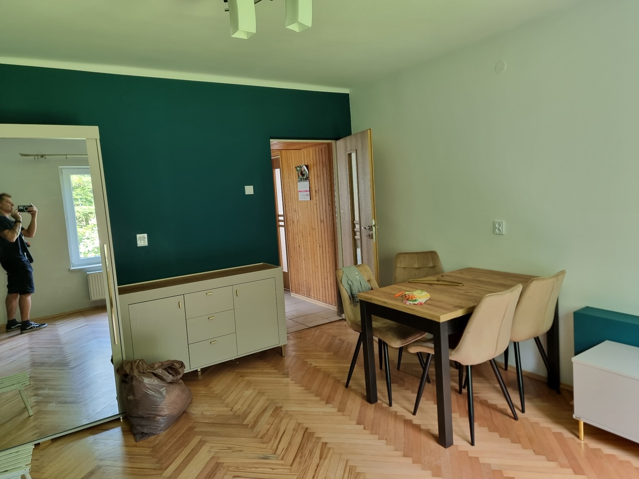 Pokój po częściowym odświeżeniu: ciemnozielona ściana, odnowiona podłoga parkietowa w jodełkę, biała komoda i stół z krzesłami; fotograf robi zdjęcie w lustrze.