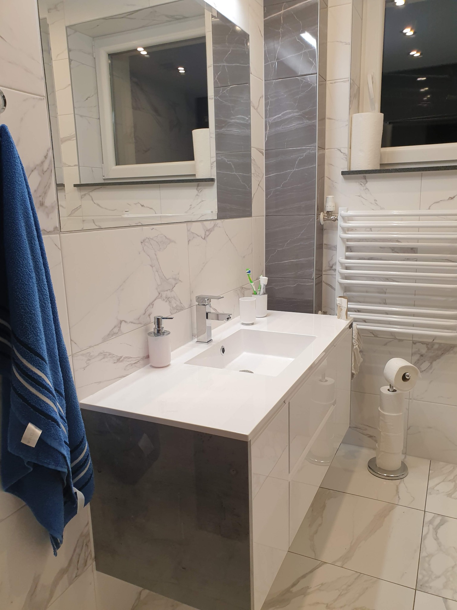 Nowoczesna łazienka z umywalką nablatową, baterią, dozownikiem mydła, grzejnikiem drabinkowym, lustrem i ręcznikiem w odcieniach bieli, szarości i błękitu, wykończona płytkami imitującymi marmur.