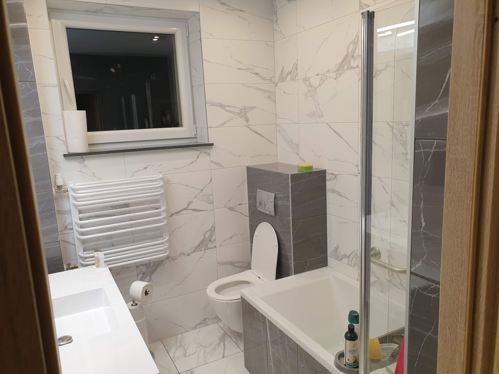 Nowoczesna łazienka z białymi płytkami imitującymi marmur, szarą obudową toalety i wanny, grzejnikiem drabinkowym oraz kabiną prysznicową ze szklanymi drzwiami.