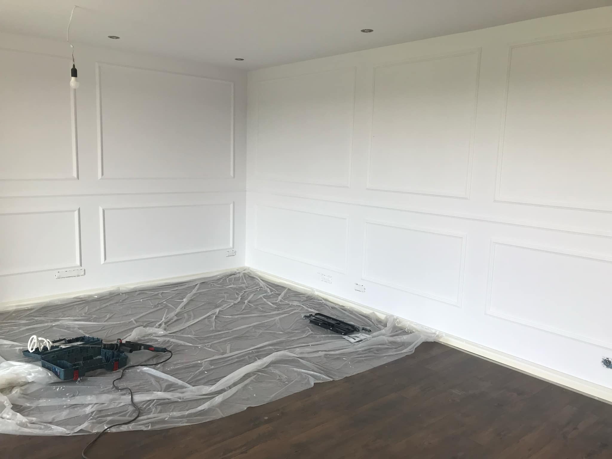 Białe ściany z dekoracyjnymi panelami w trakcie przygotowań do malowania, podłoga zabezpieczona folią malarską, widoczny otwarty kufer z narzędziami malarskimi i wisząca żarówka.