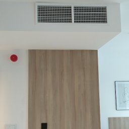 Mikołaj Skałecki Remont w Twoim stylu - Nowoczesne wnętrze: gładkie, białe ściany z kratką wentylacyjną, drewniana okładzina ściany z czarnym uchwytem, w tle fragment sypialni z łóżkiem i obrazem.