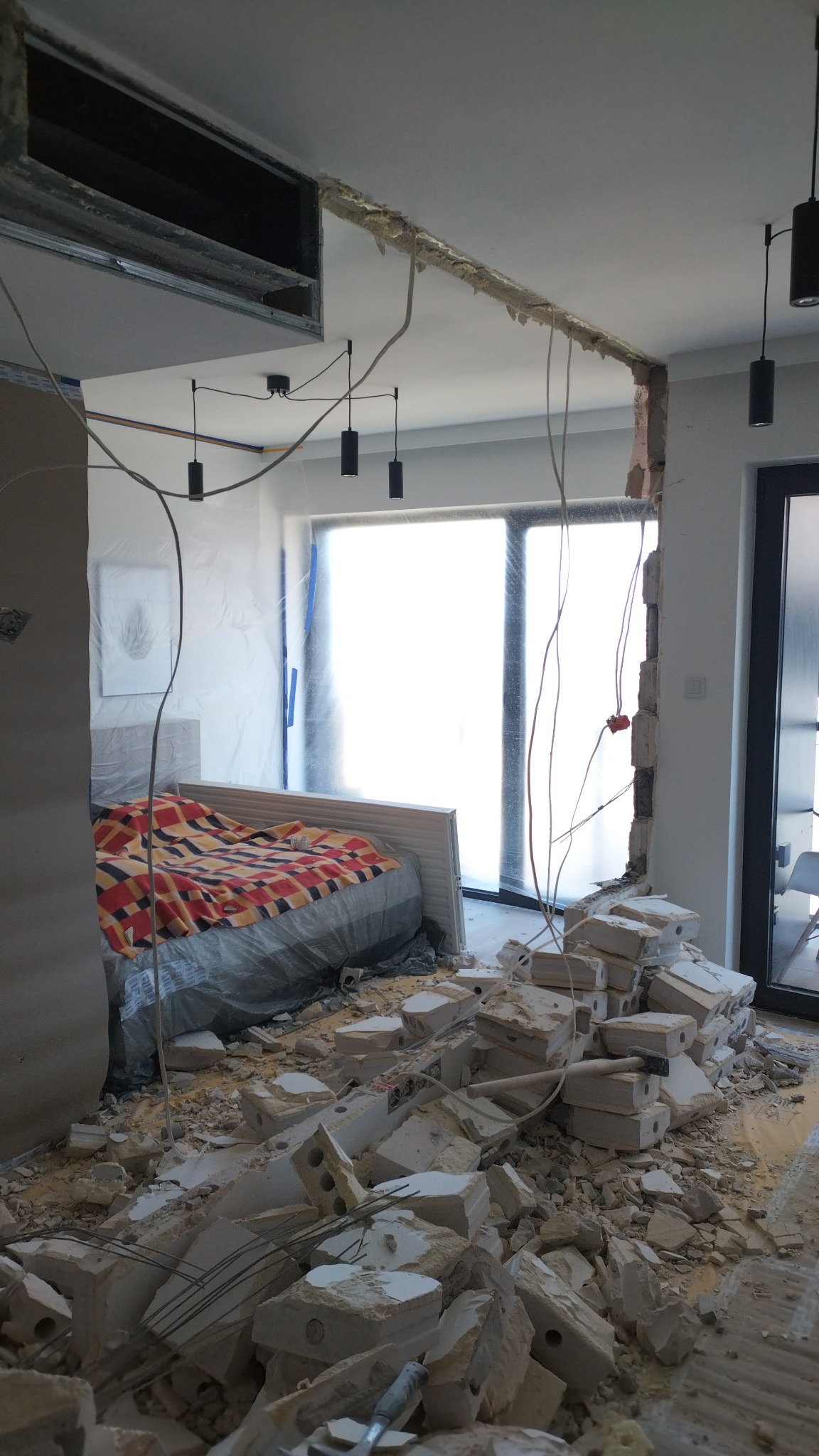 Demontaż ściany działowej w pokoju, gruz na podłodze, zabezpieczone meble folią, widoczne przewody elektryczne i elementy oświetlenia. Remont w toku.