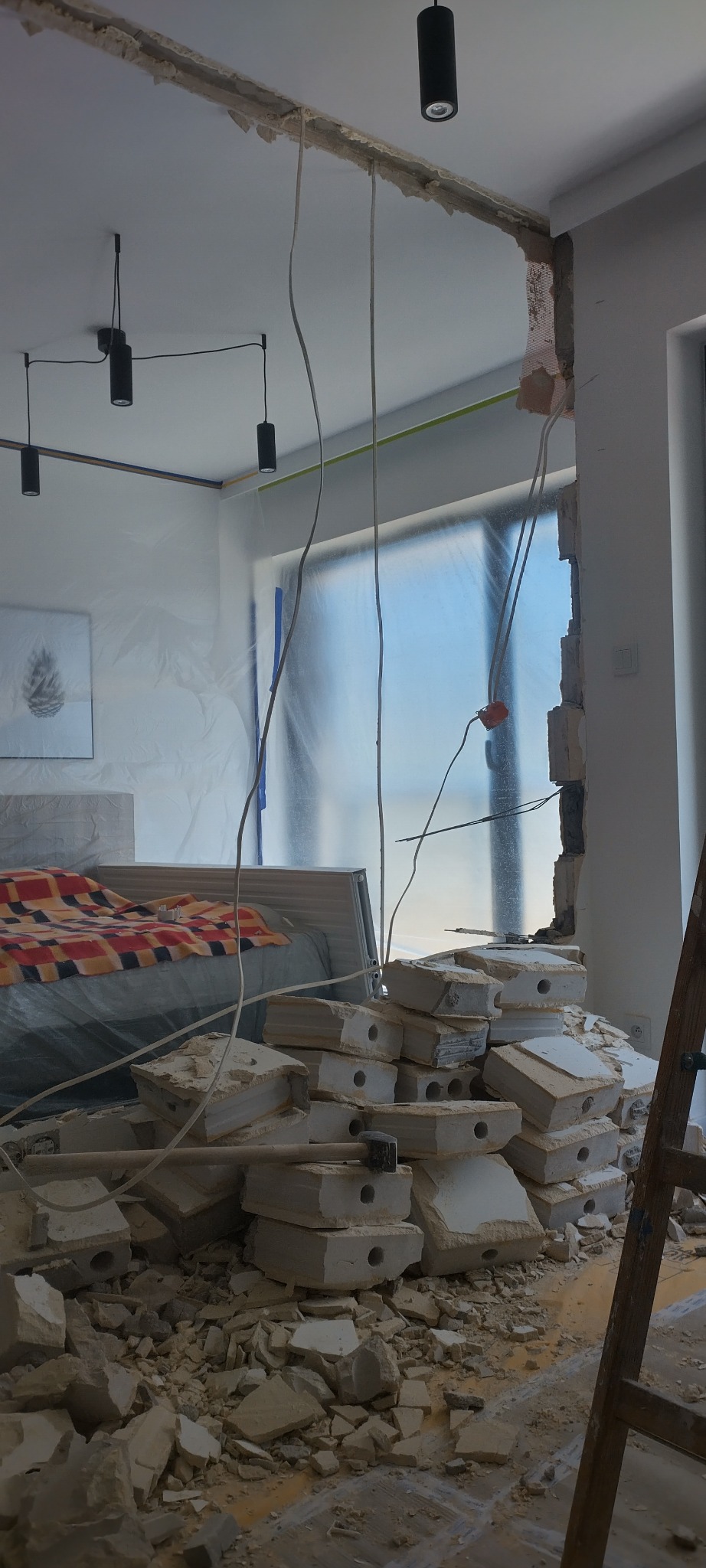 Demontaż ściany w pokoju z zabezpieczonym folią oknem i meblami. Widoczny stos gruzu i młotek. Odsłonięte kable elektryczne.
