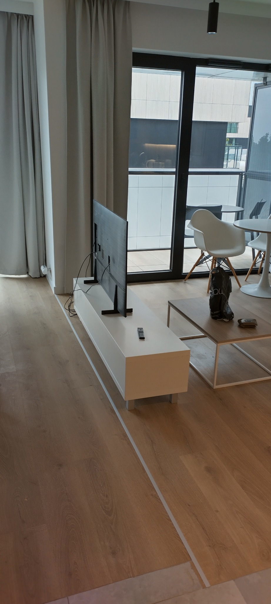 Jasne panele podłogowe w nowoczesnym salonie. Biała listwa przypodłogowa oddziela podłogę od ściany. Widoczny fragment szafki RTV, okna balkonowe i krzesła.