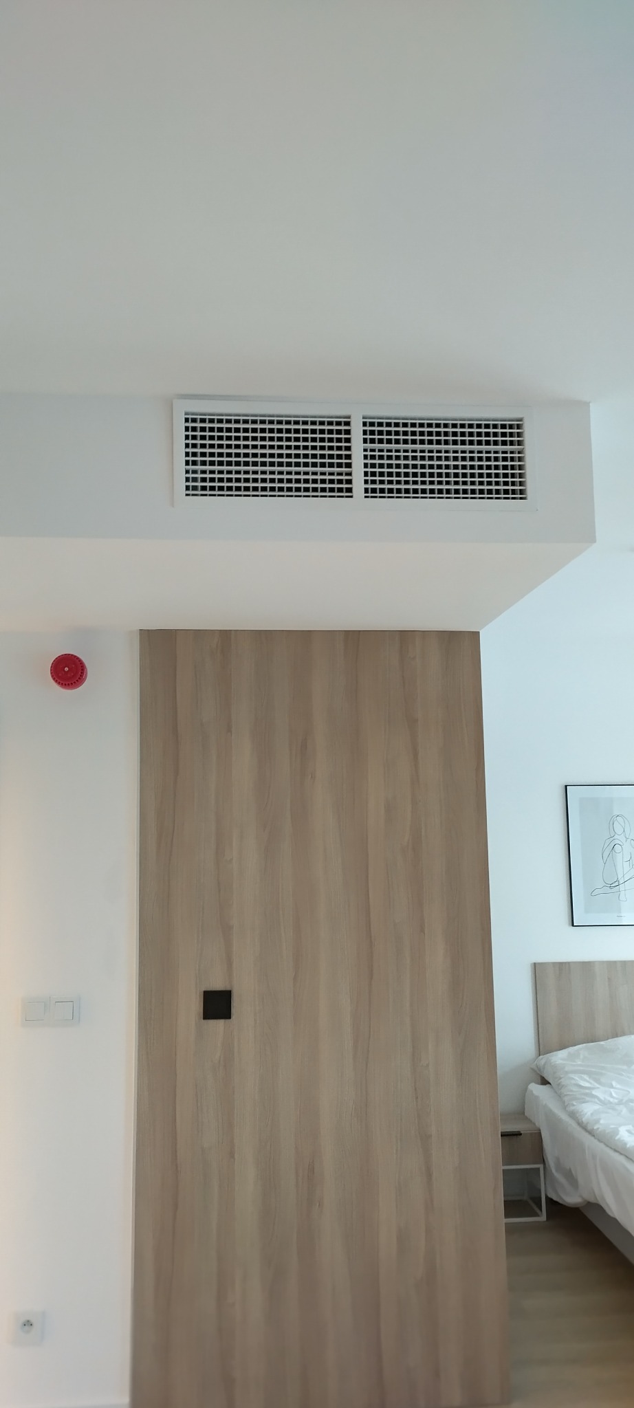 Nowoczesne wnętrze: gładkie, białe ściany z kratką wentylacyjną, drewniana okładzina ściany z czarnym uchwytem, w tle fragment sypialni z łóżkiem i obrazem.