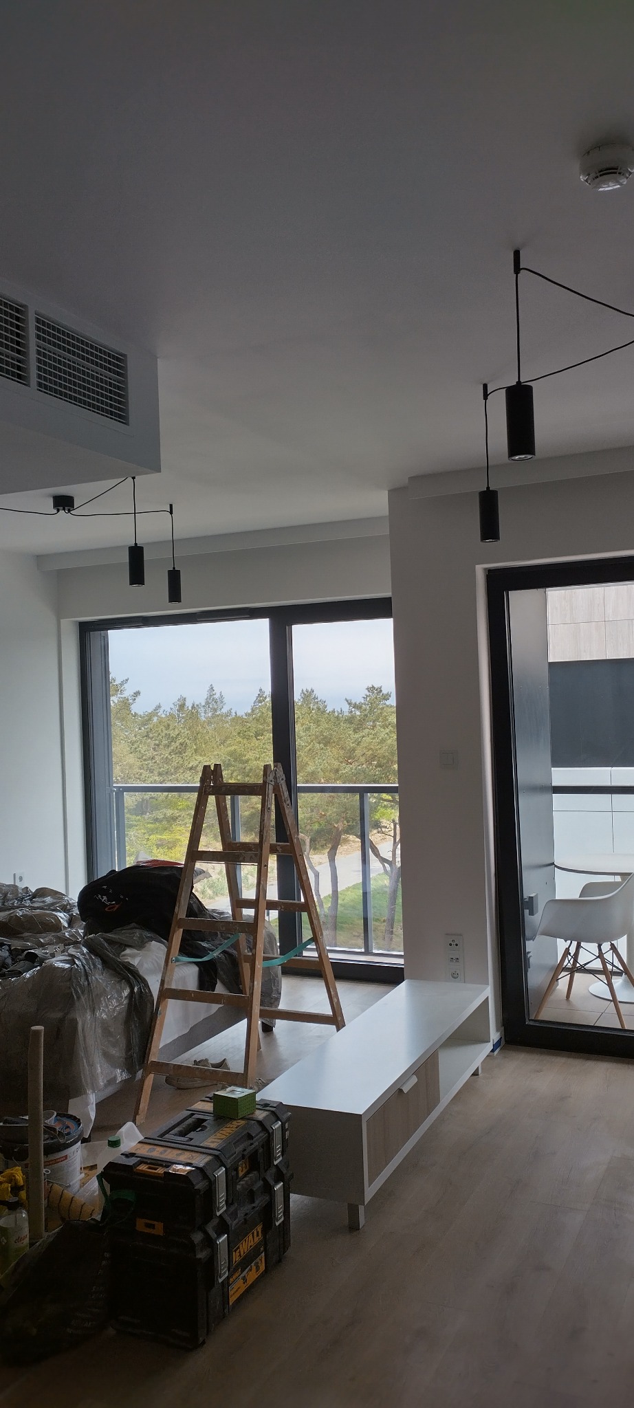 Wnętrze w trakcie remontu: drabina oparta o okno z widokiem na drzewa, skrzynka narzędziowa na podłodze, meble zabezpieczone folią malarską. Widoczne elementy oświetlenia.