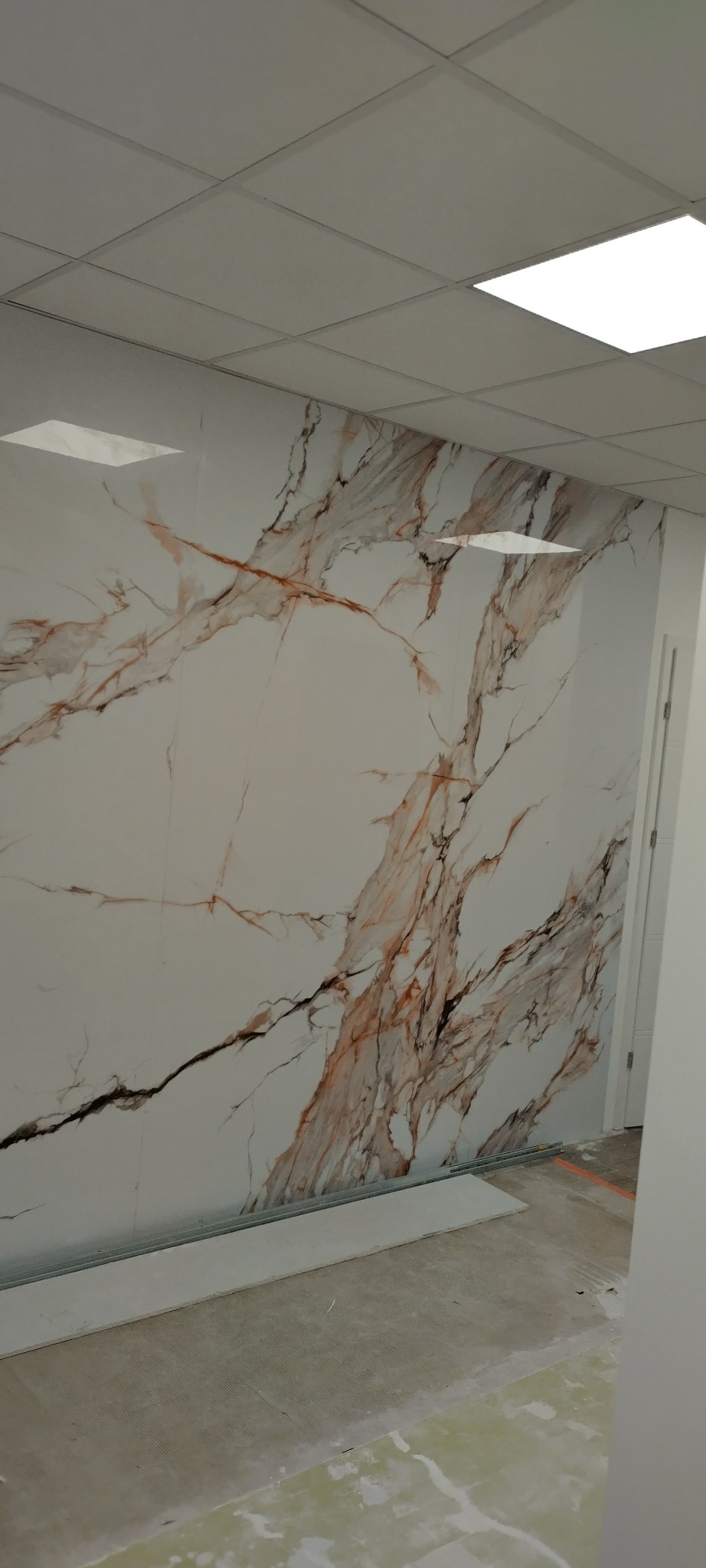 Ściana wykończona płytami imitującymi marmur z brązowymi żyłami, widoczny sufit kasetonowy i fragment podłogi w trakcie prac wykończeniowych.