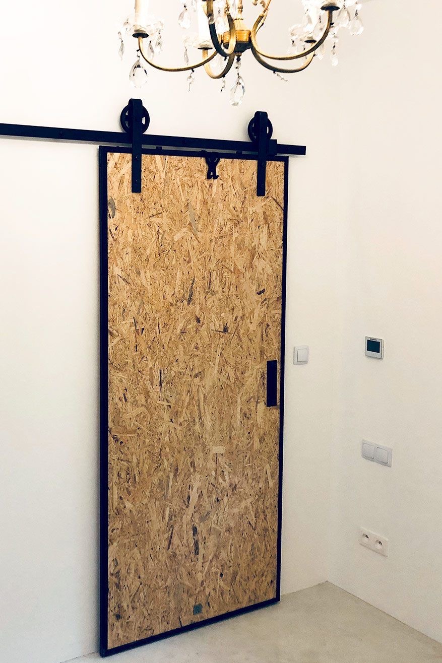 Suwane drzwi z płyty OSB na czarnej szynie, zawieszone w jasnym wnętrzu pod kryształowym żyrandolem.