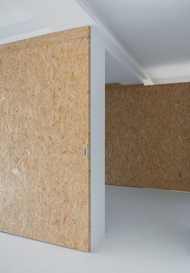 Wnętrze z surowymi ścianami wykończonymi płytami OSB, białe podłogi i sufit, minimalistyczny design z ukrytymi drzwiami przesuwnymi z prostokątnym uchwytem.