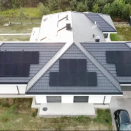 Widok z drona na dom jednorodzinny z ciemną dachówką i zamontowanymi panelami słonecznymi na kilku połaciach dachu; obok domu stoi czarny samochód.