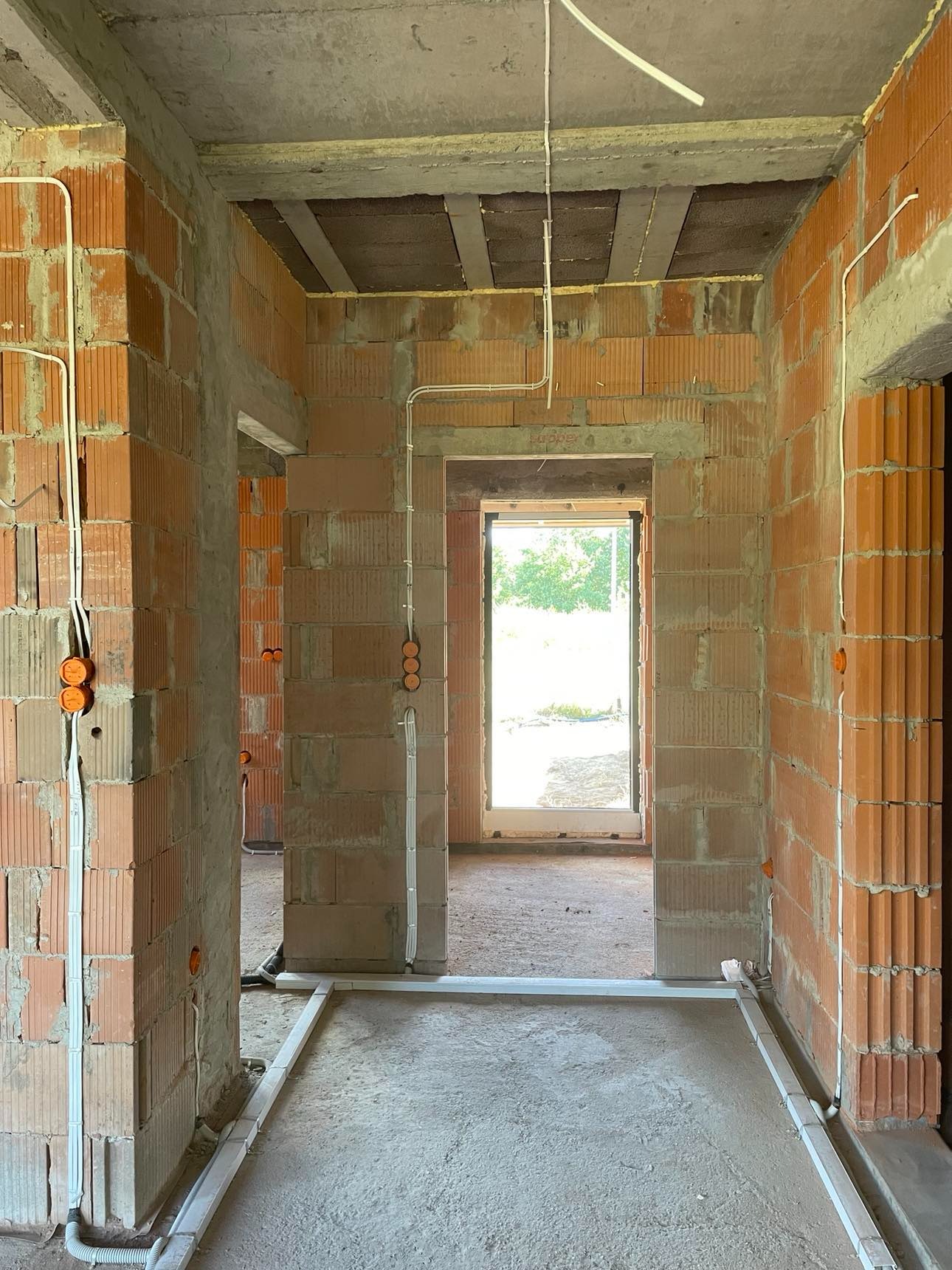 Wnętrze budynku w stanie surowym otwartym, widoczne rozprowadzone przewody elektryczne w peszlach na ścianach z cegły oraz pod sufitem, przygotowane puszki instalacyjne.