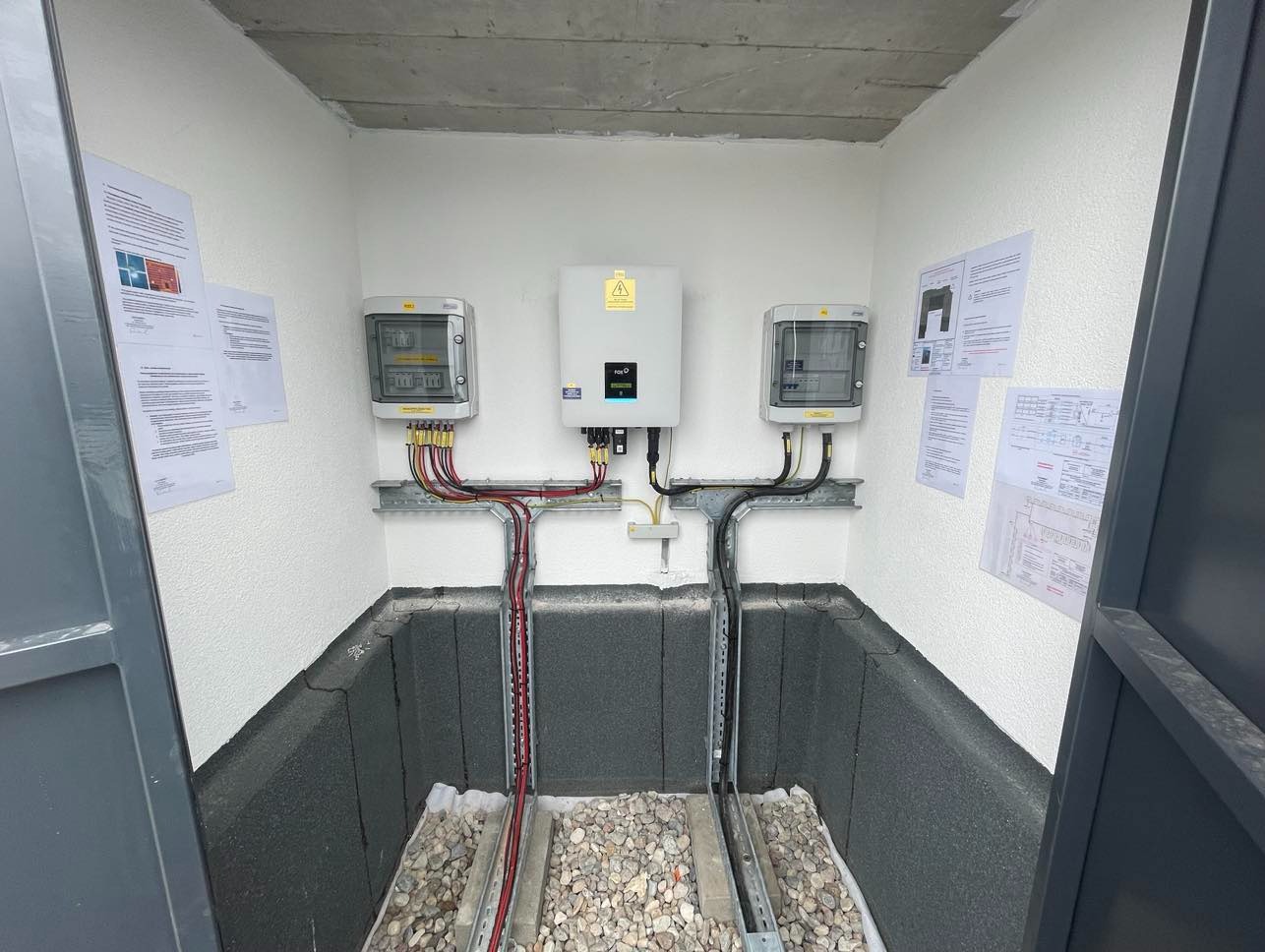 Wewnętrzna instalacja elektryczna w pomieszczeniu technicznym, z widocznymi dwoma rozdzielnicami, inwerterem centralnym z ostrzeżeniem o napięciu, oraz okablowaniem. Na ścianach zawieszone...