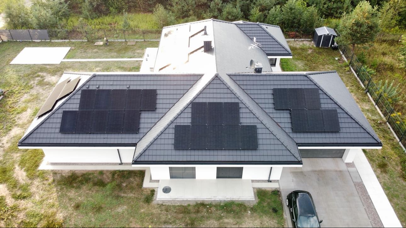 Widok z drona na dom jednorodzinny z ciemną dachówką i zamontowanymi panelami słonecznymi na kilku połaciach dachu; obok domu stoi czarny samochód.
