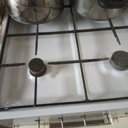 Robert hibner - Brudna, biała kuchenka gazowa z dwoma garnkami na ruszcie, widoczne przypalenia i zabrudzenia na płycie i palnikach.