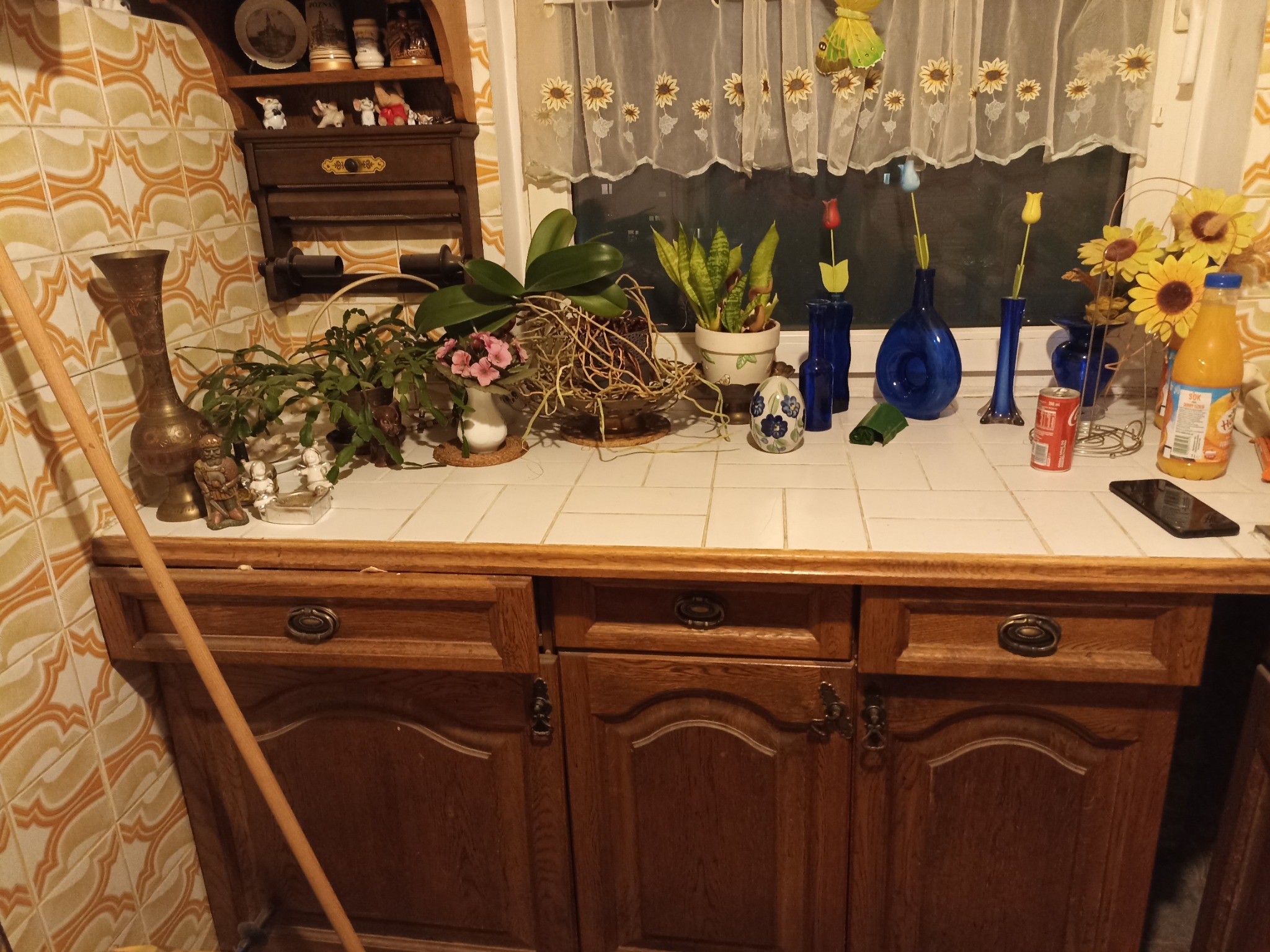 Kuchnia w stylu retro z drewnianymi szafkami, blatem wyłożonym kafelkami i dekoracjami: kwiaty w wazonach, ozdobne figurki, butelki z sokiem i ściereczka z motywem słoneczników w oknie.