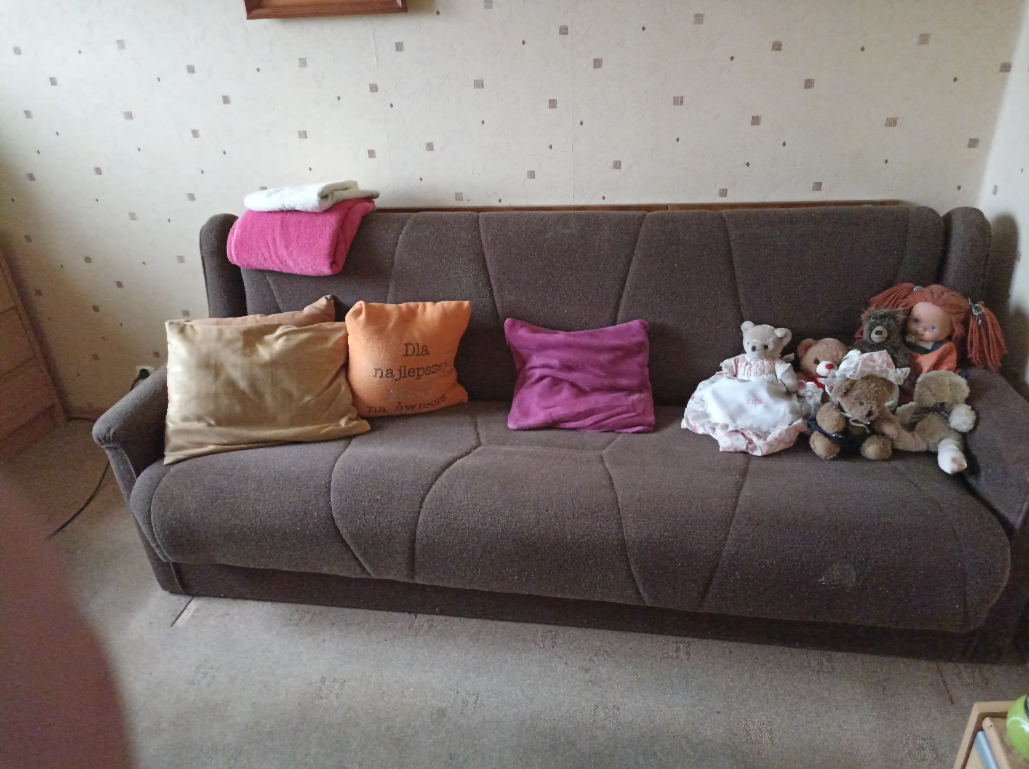 Brązowa sofa z poduszkami i pluszowymi zabawkami w pokoju z wzorzystą tapetą. Poduszka z napisem 'Dla najlepszej'.