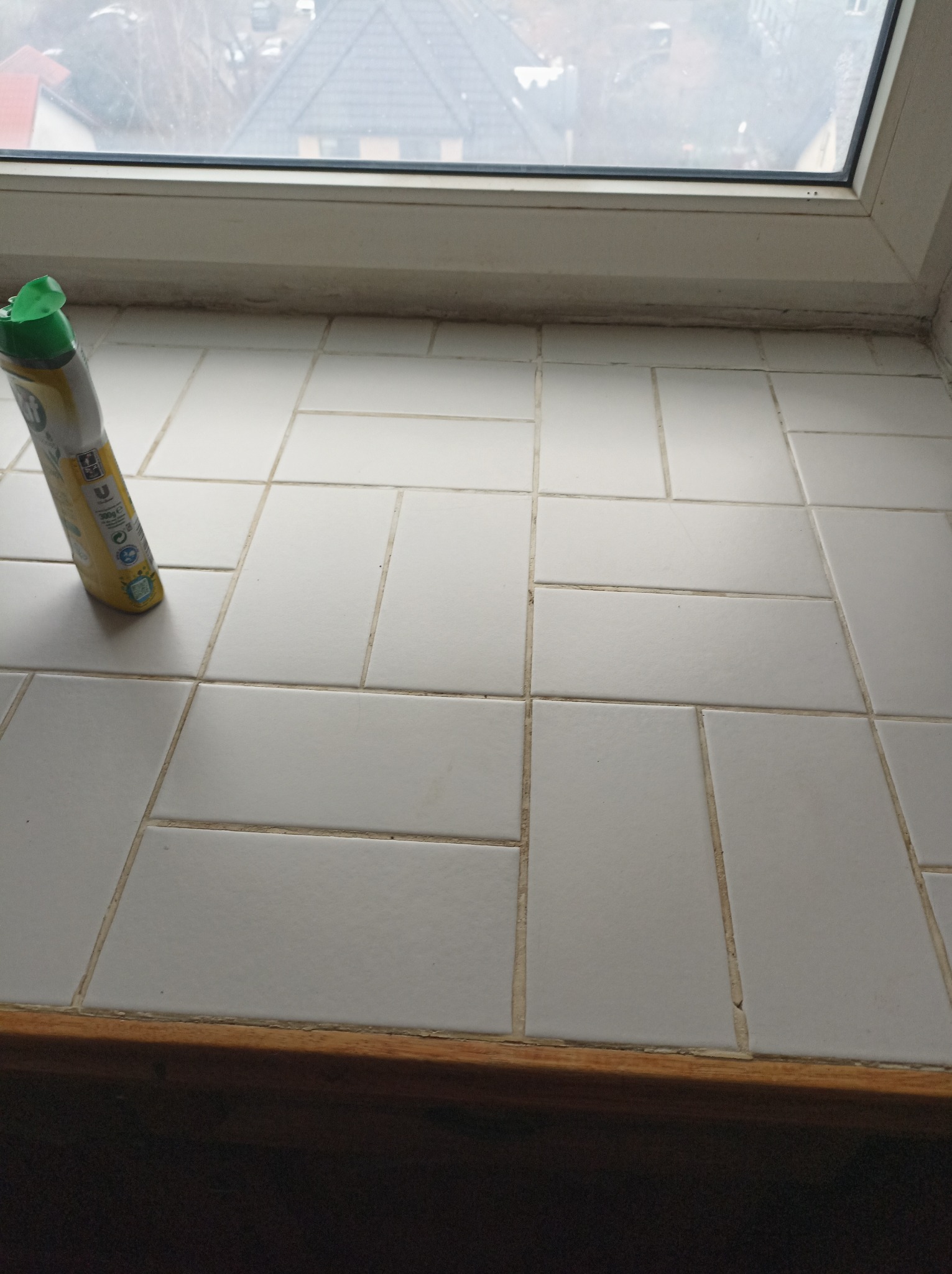 Butelka z płynem czyszczącym leży na białym, ceramicznym parapecie okiennym z widocznymi fugami, okno z widokiem na miasto w tle.