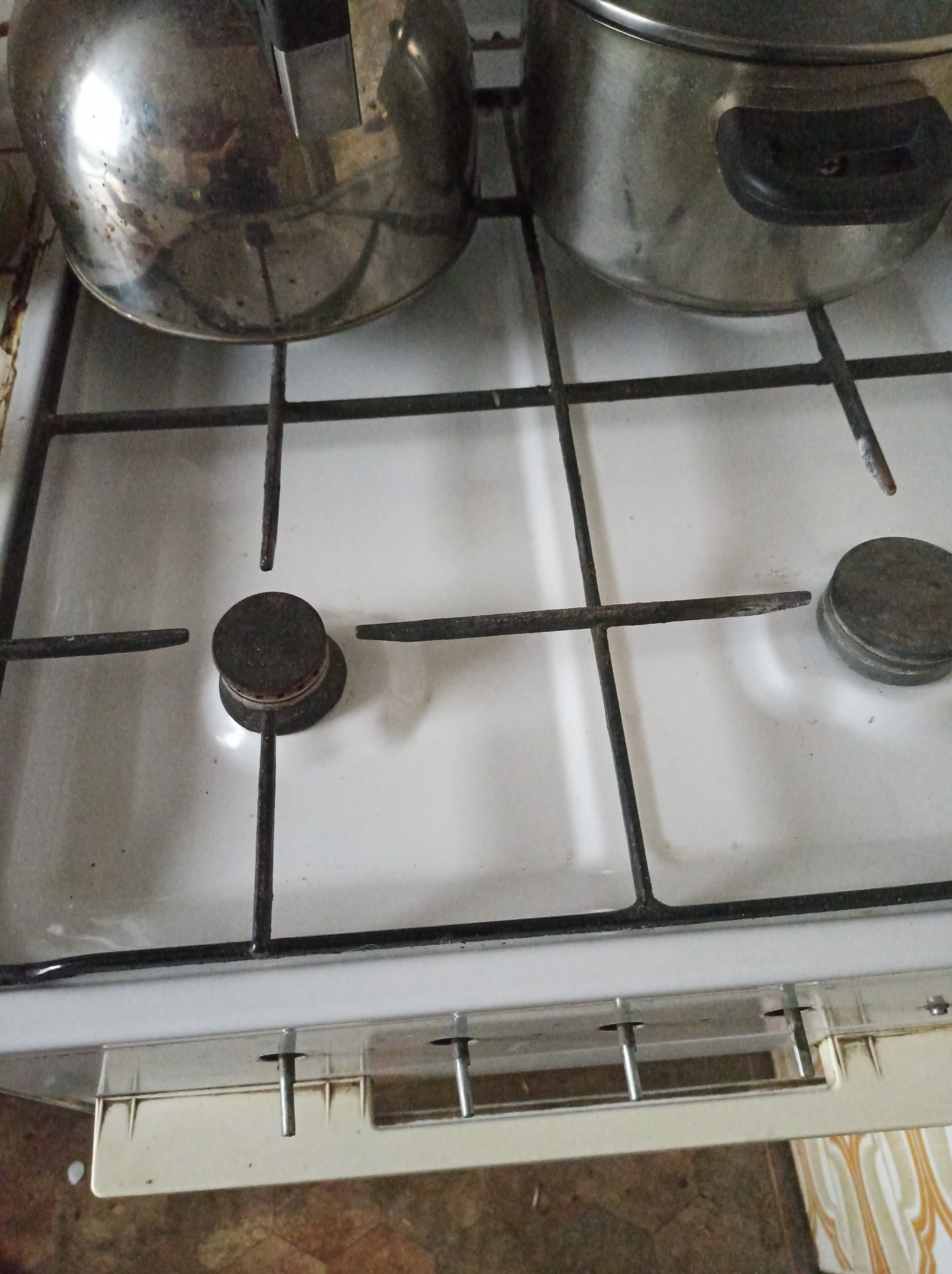 Brudna, biała kuchenka gazowa z dwoma garnkami na ruszcie, widoczne przypalenia i zabrudzenia na płycie i palnikach.