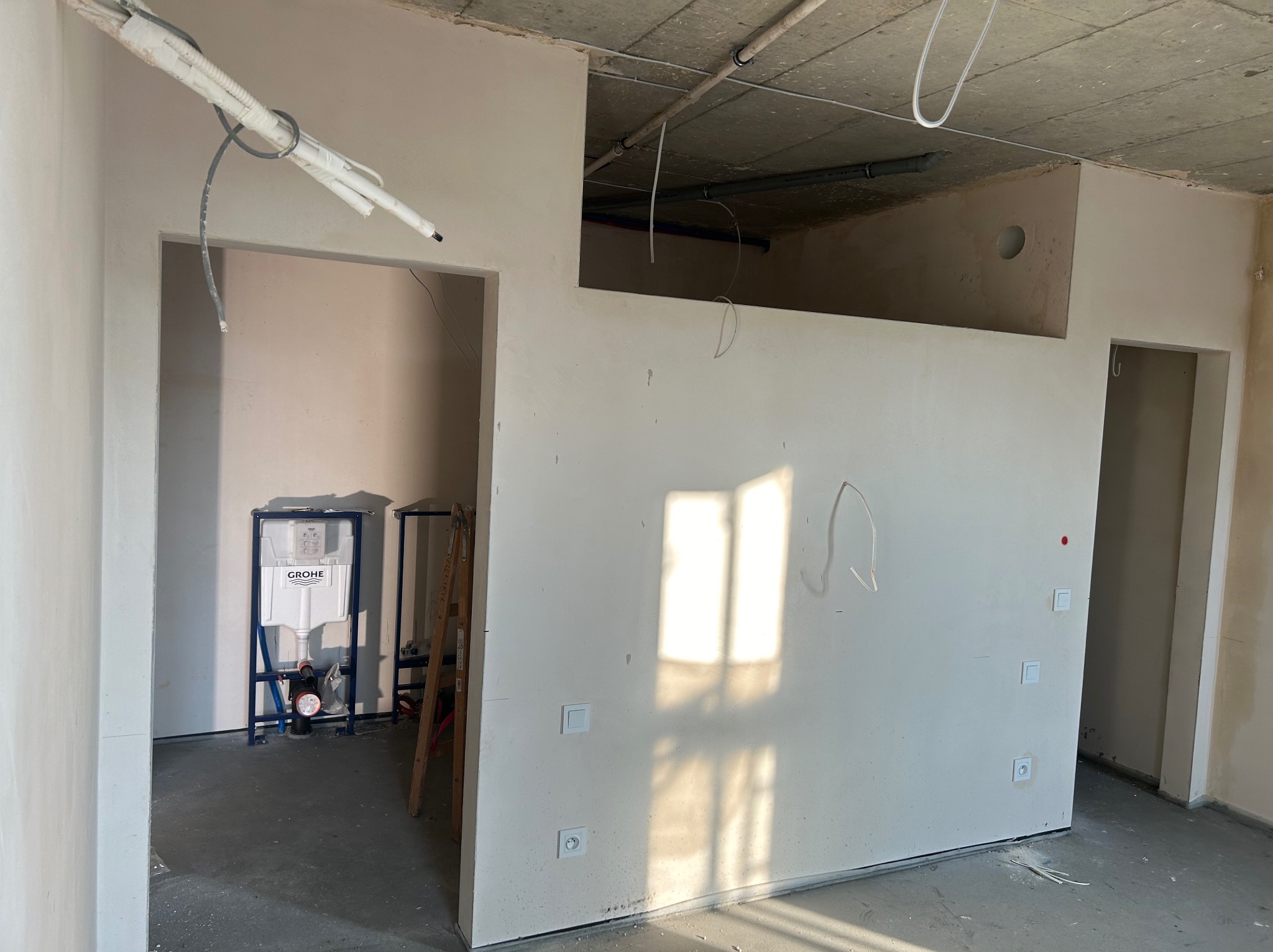 Wnętrze w trakcie prac wykończeniowych: ściany pokryte świeżym, jasnym tynkiem, widoczne instalacje elektryczne, fragment instalacji sanitarnej oraz drabina oparta o ścianę.