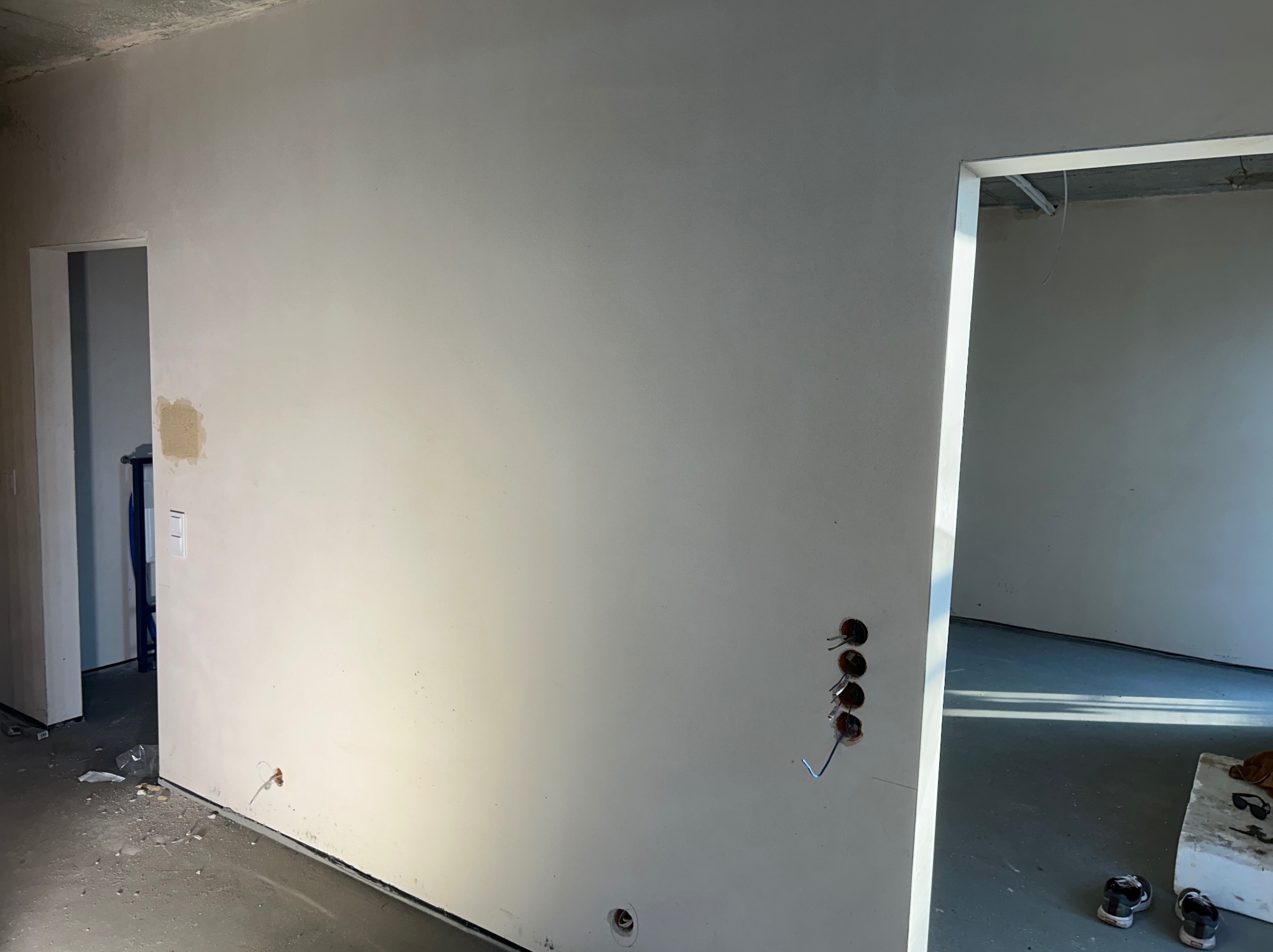 Świeżo otynkowana ściana w surowym wnętrzu z widocznymi otworami na gniazdka elektryczne i wyprowadzonymi przewodami, z fragmentem niedokładności w rogu przy przejściu do innego pomieszczenia.