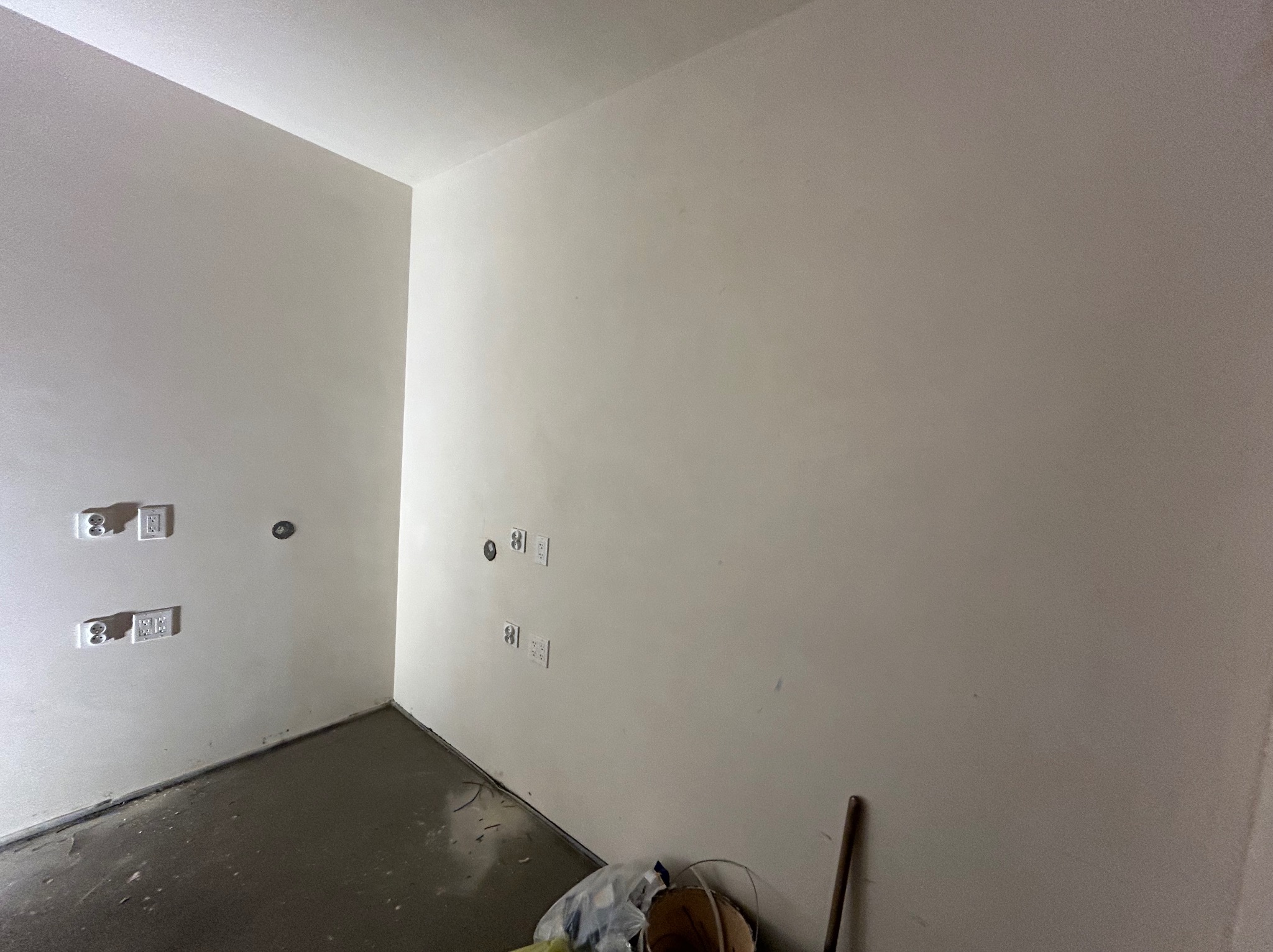 Wnętrze po tynkowaniu maszynowym: gładkie, jasne ściany z widocznymi gniazdkami elektrycznymi i niedoskonałościami posadzki, widoczny fragment wiadra i kija do mieszania zaprawy w rogu pomieszczenia.