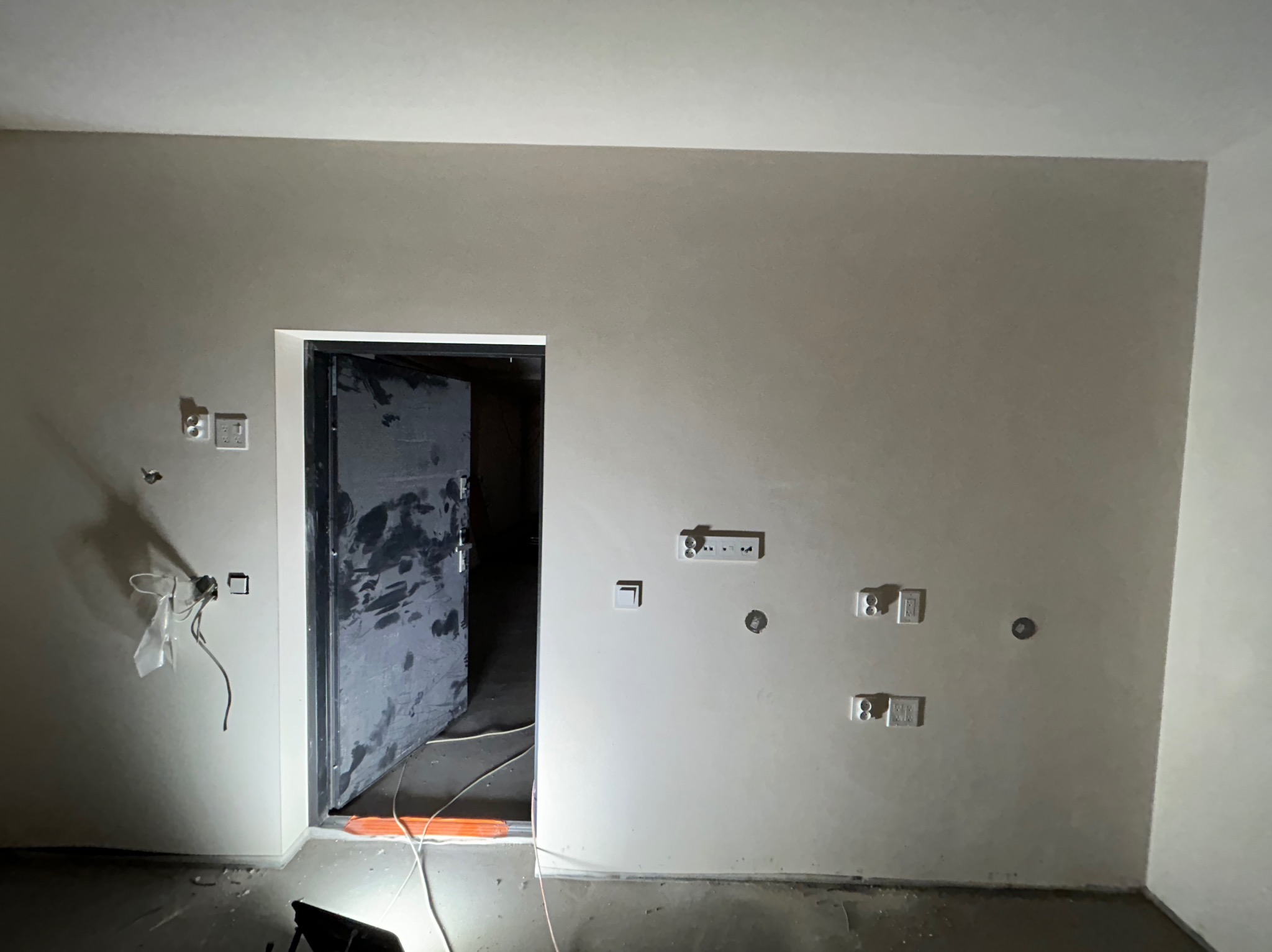 Świeżo otynkowane ściany w pomieszczeniu z otwartymi drzwiami, widoczne gniazdka elektryczne i wystające przewody, podłoga częściowo zabezpieczona folią.