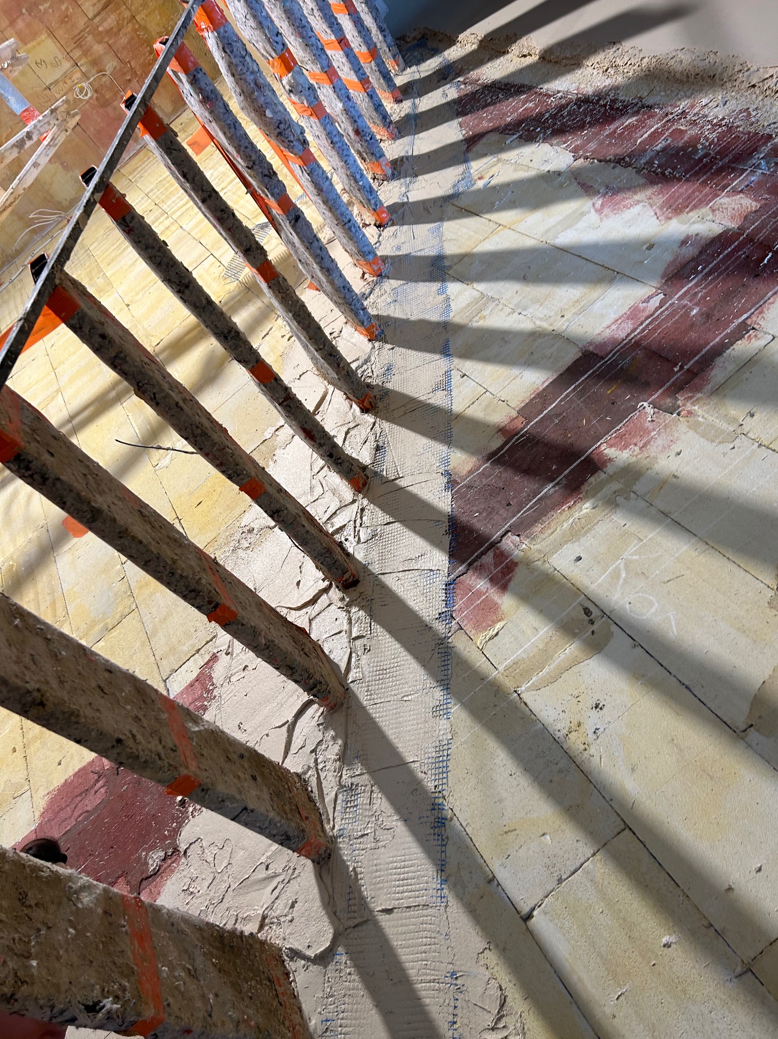 Przygotowanie podłoża pod tynki: fragment ściany z naniesioną siatką wzmacniającą, widoczne pionowe elementy rusztowania z pomarańczowymi oznaczeniami, rzucające wyraźny cień na podłoże.