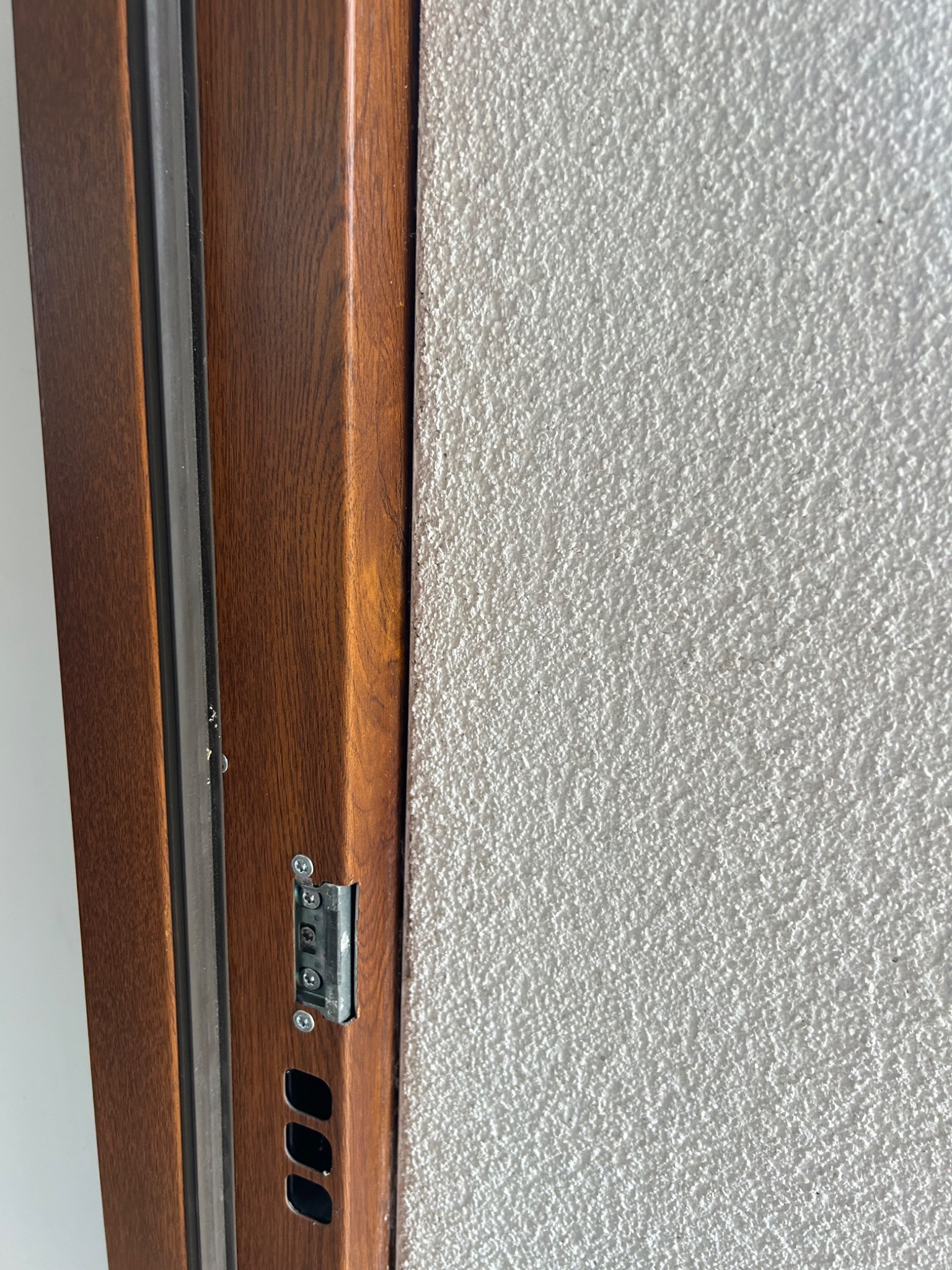 Fragment ściany z dekoracyjną, białą strukturą tynku obok brązowej futryny drzwiowej z widocznym metalowym elementem mocującym.