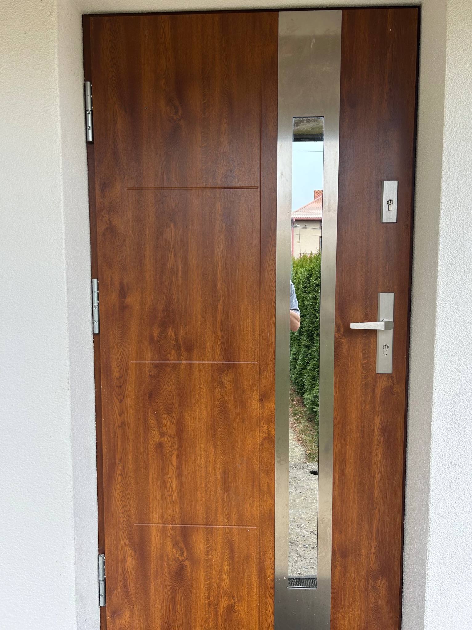 Brązowe drzwi wejściowe z metalową wstawką i odblaskowym elementem, widoczne zawiasy i klamka na tle białego muru.