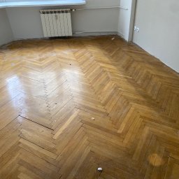 Renowacja schodów - cyklinowanie podłóg - Parkiet ułożony w jodełkę przed renowacją, widoczne uszkodzenia i pozostałości po pracach remontowych przy ścianach.