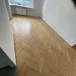 Renowacja schodów - cyklinowanie podłóg - Parkiet ułożony w jodełkę po renowacji w jasnym pokoju z oknem i grzejnikiem, widoczne ślady po listwach przypodłogowych.