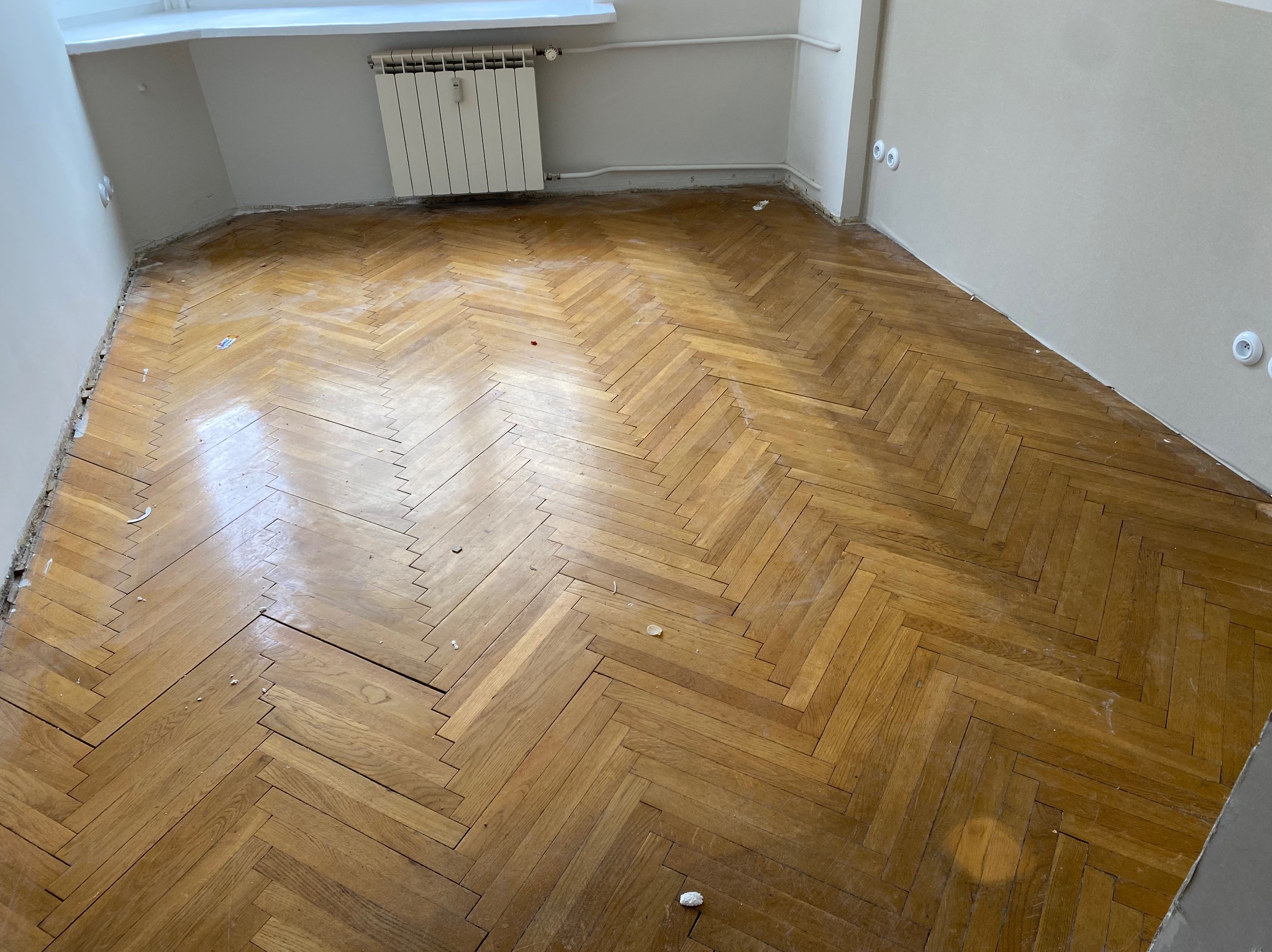 Parkiet ułożony w jodełkę przed renowacją, widoczne uszkodzenia i pozostałości po pracach remontowych przy ścianach.