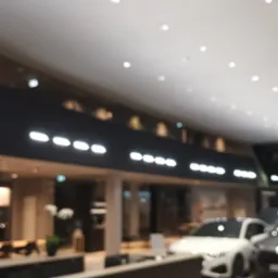 Rozmyte wnętrze salonu samochodowego z widocznymi samochodami, oświetleniem punktowym i elementami wystroju.