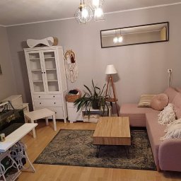 Expert-Bud - Jasnoszary salon z pudrowo różową sofą narożną, drewnianym stolikiem kawowym na metalowych nóżkach, białym regałem z przeszkleniami, akwarium na stole ze starą podstawą maszyny do szycia i dywanem...