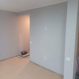 Expert-Bud - Jasnoszary pokój po malowaniu w Aleksandrowie Łódzkim, z drewnianą podłogą i skrzynką narzędziową w tle, widoczne gniazdka elektryczne i włączniki.