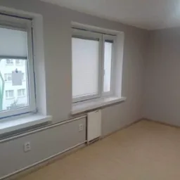 Pomalowane na szaro wnętrze pokoju z dwoma oknami z roletami, jasna podłoga, widoczne gniazdka elektryczne na ścianie pod oknem.