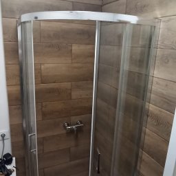 Expert-Bud - Narożna kabina prysznicowa z przesuwanymi drzwiami, wyłożona płytkami imitującymi drewno, z widoczną baterią i ręcznikiem w brodziku. Z lewej strony pralka i narzędzia.