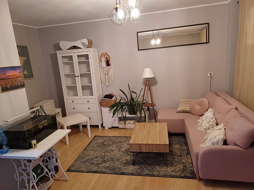 Jasnoszary salon z pudrowo różową sofą narożną, drewnianym stolikiem kawowym na metalowych nóżkach, białym regałem z przeszkleniami, akwarium na stole ze starą podstawą maszyny do szycia i dywanem...