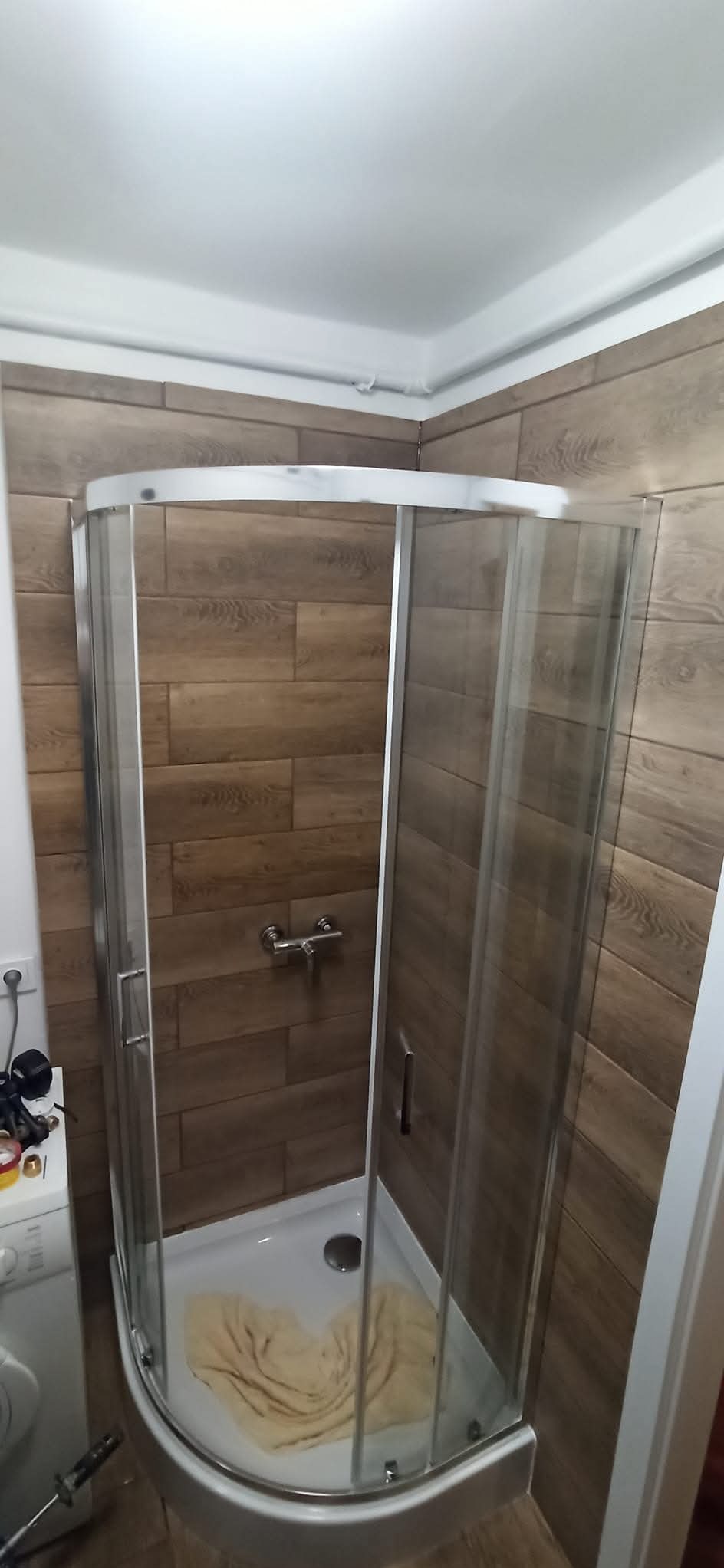 Narożna kabina prysznicowa z przesuwnymi drzwiami, zamontowana w łazience wyłożonej brązowymi płytkami imitującymi drewno, widoczny biały brodzik z ręcznikiem w środku oraz bateria.